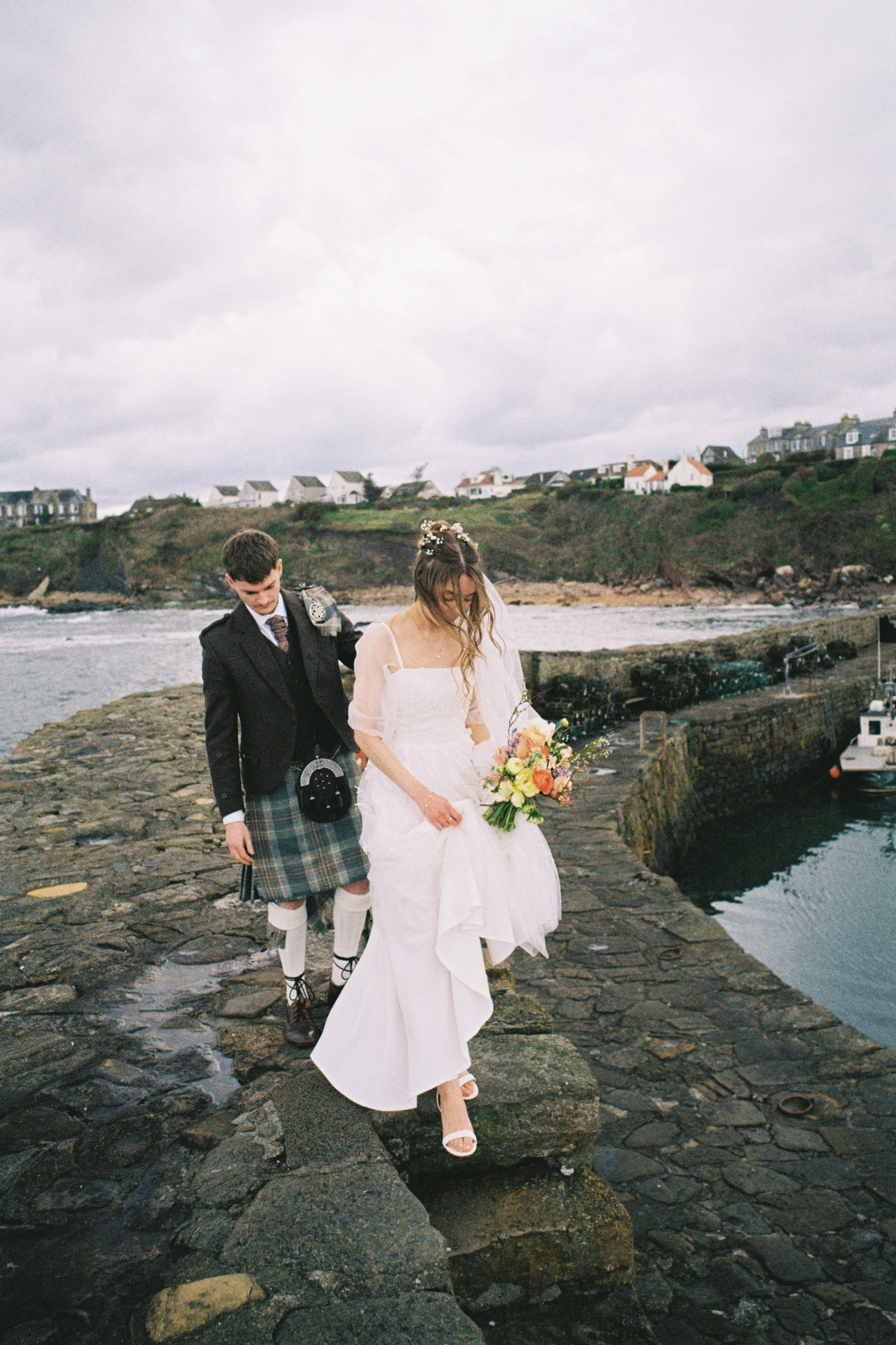 A bride in a white dress holding a bouquet walking on rocks near water, with a groom in Scottish attire behind her, on a cloudy day with houses on a hill in the background.