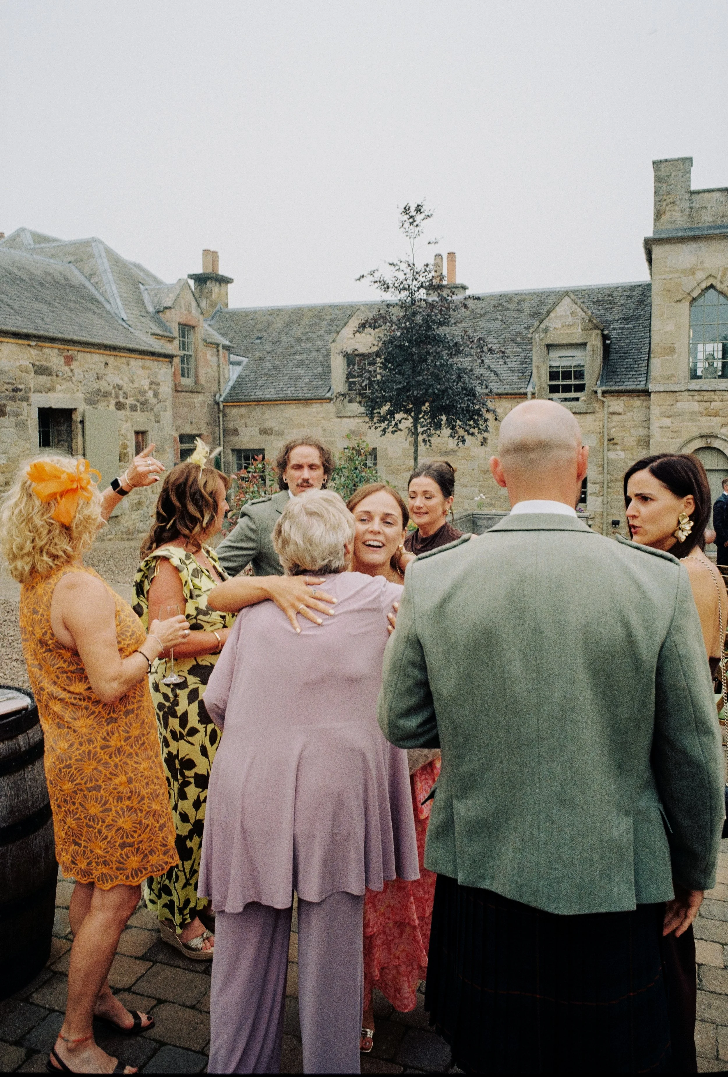 A group of people socializing outdoors in front of a stone building, dressed in colorful, semi-formal attire, with women wearing dresses and men in suits.