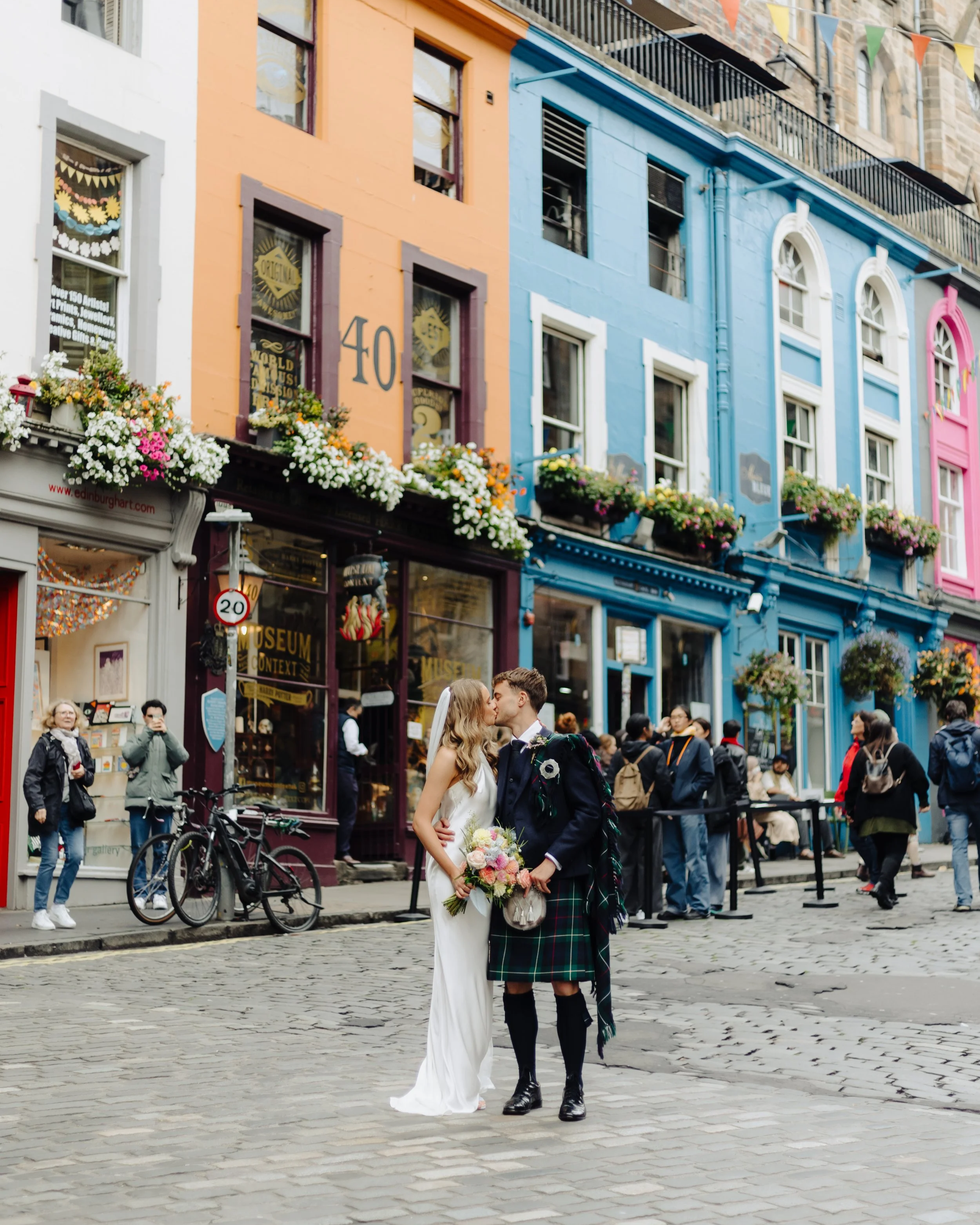 A bride and groom sharing a kiss on a cobblestone street in front of a colorful building with flowers and spectators watching.