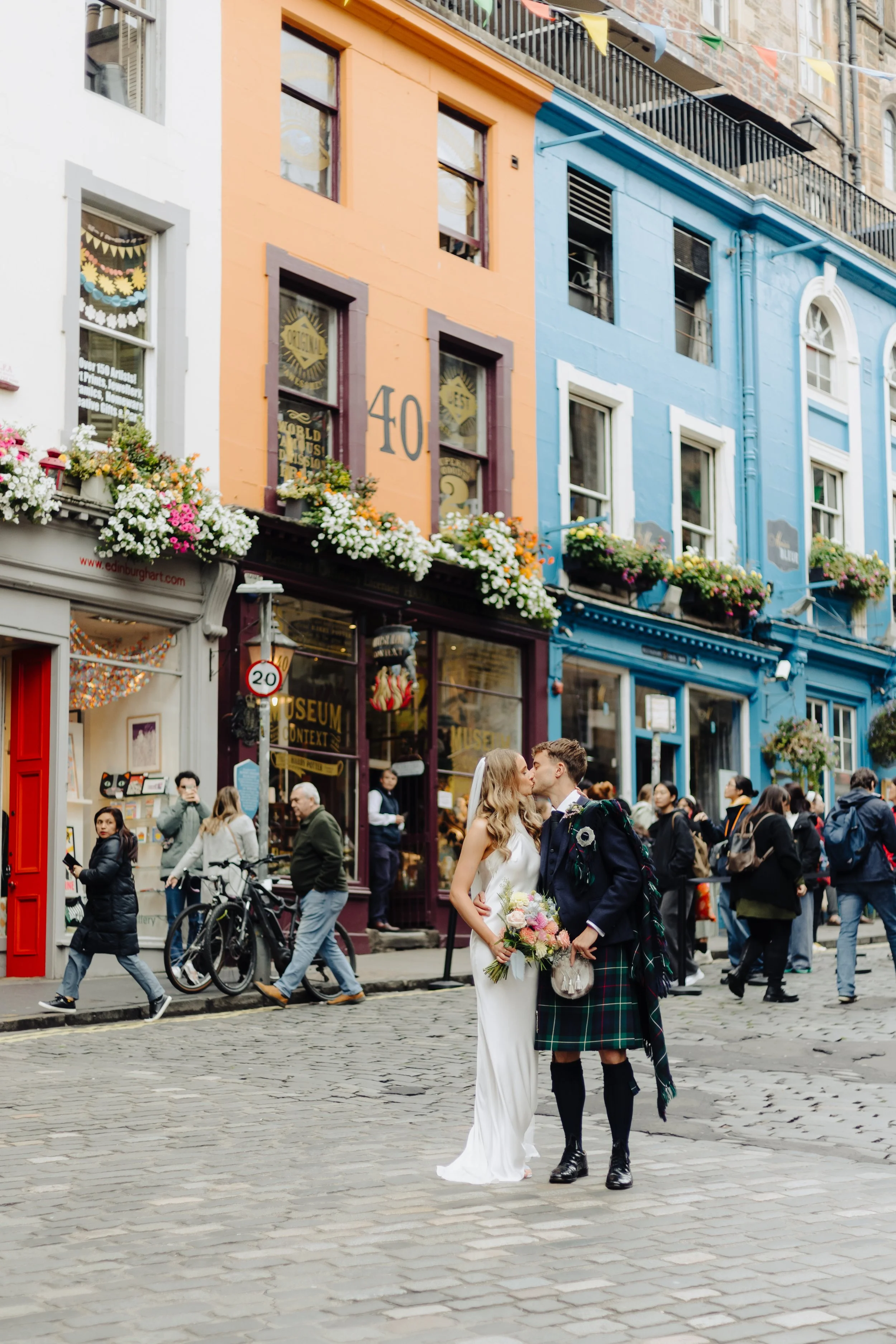 A bride and groom sharing a kiss in a busy city street with colorful buildings, flower boxes, and pedestrians, including people taking photos and bicycles.