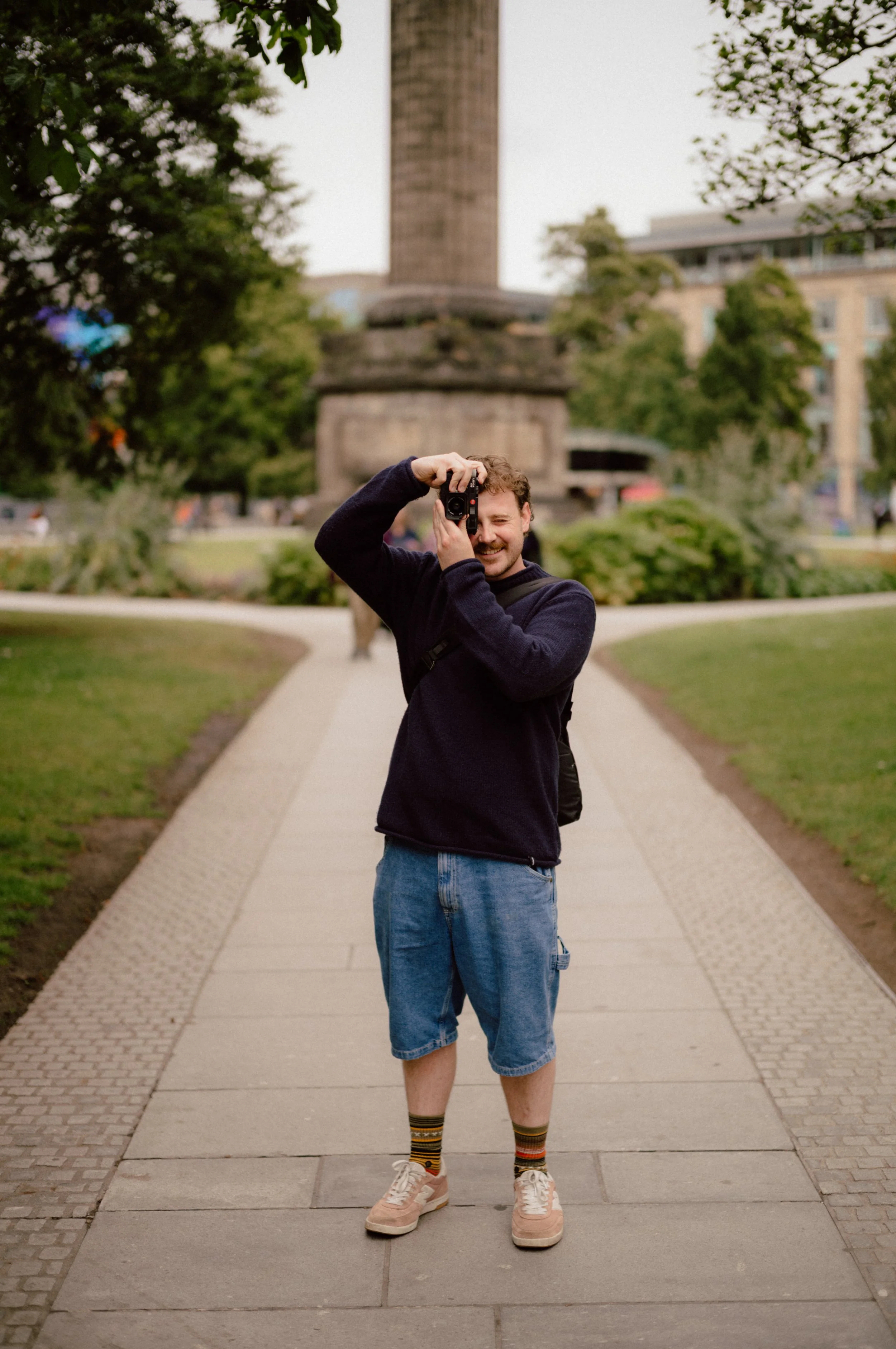 A man with curly hair and a mustache is standing on a pathway in a park, taking a photo with a camera. He is wearing a navy blue sweater, denim shorts, colorful striped socks, and pink sneakers. There are trees and a monument in the background.
