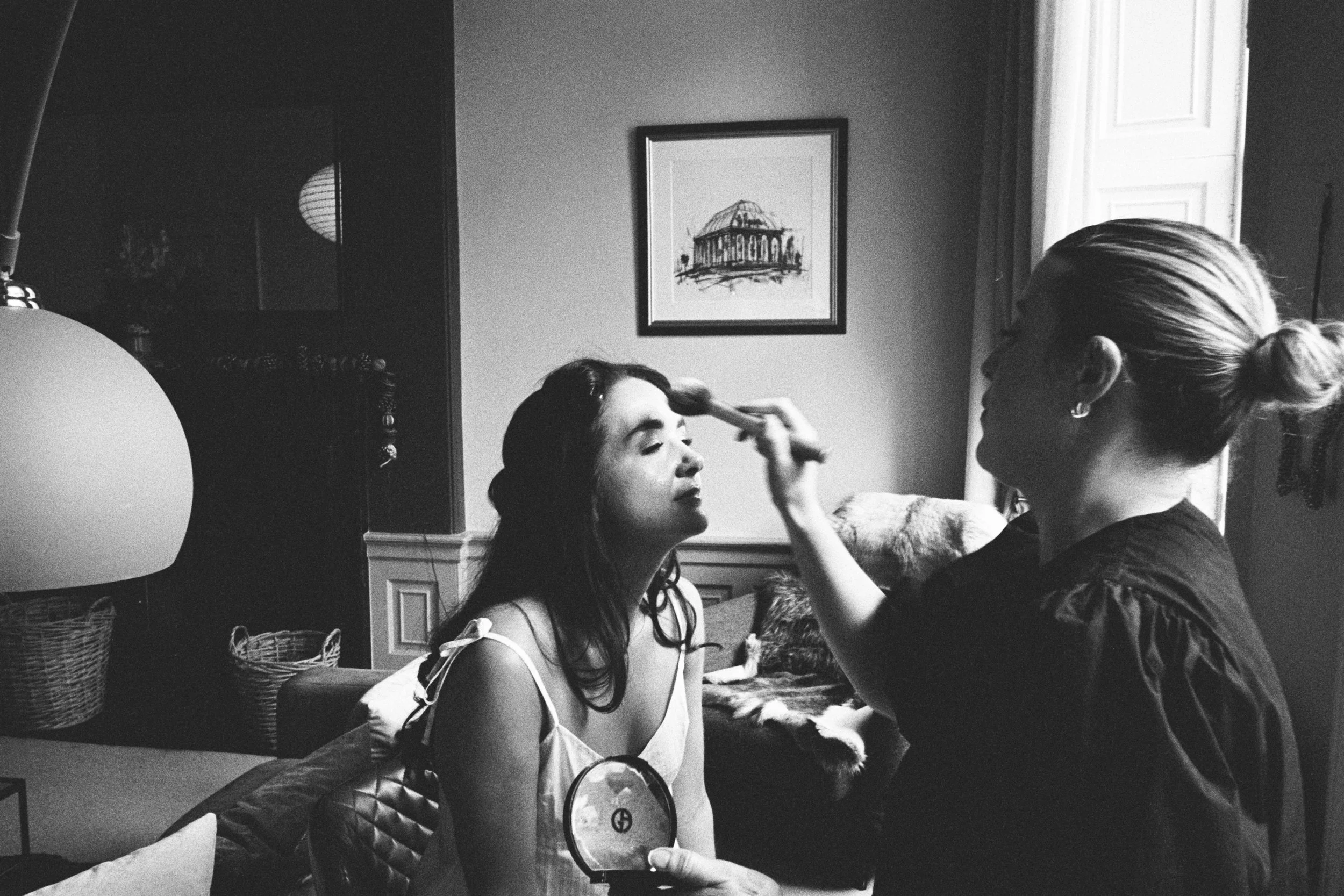A woman receives makeup application from a makeup artist. The woman is sitting with her eyes closed, and the artist is working on her face in a room with framed artwork and a large window.