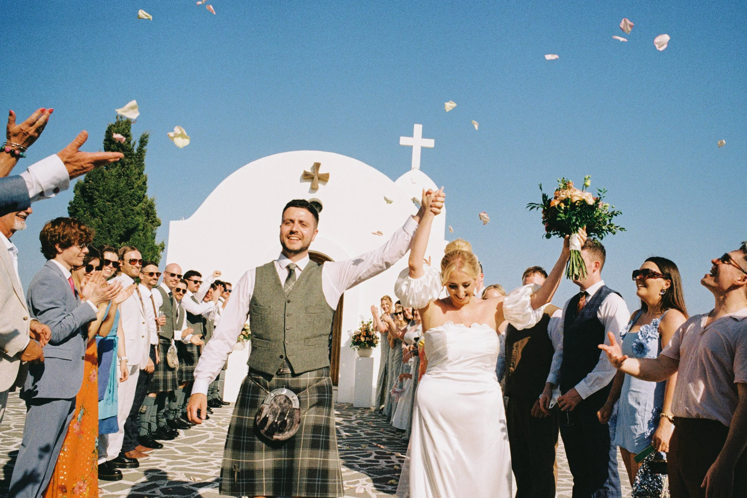 A wedding celebration outside a white chapel with a cross on top, with guests clapping and throwing flower petals, and a couple dancing in the center.