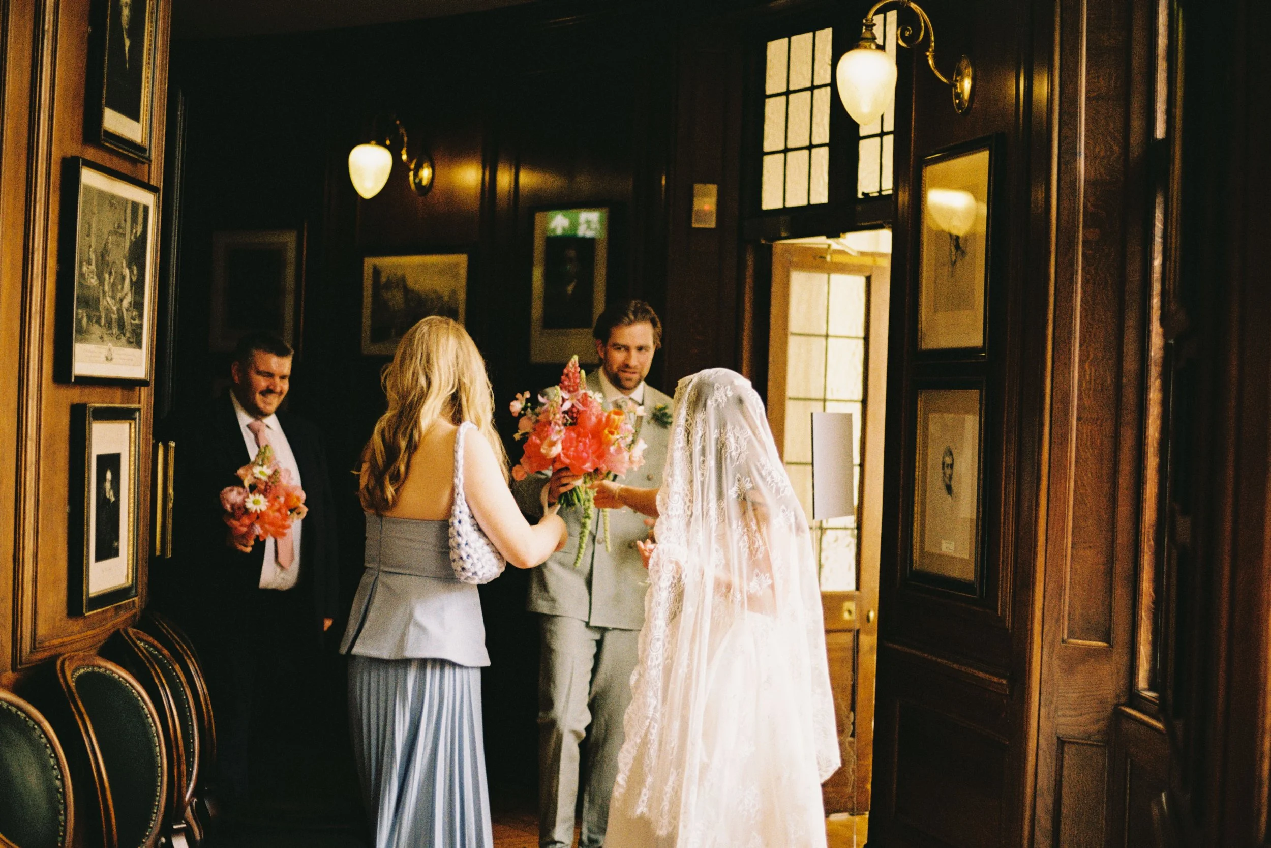 People in formal attire, likely at a wedding, are gathered in a warmly lit, wooden-paneled room with framed artwork on the walls. A bride in a white lace wedding gown and veil holds a bouquet of pink flowers, while others, including a man in a suit, observe.