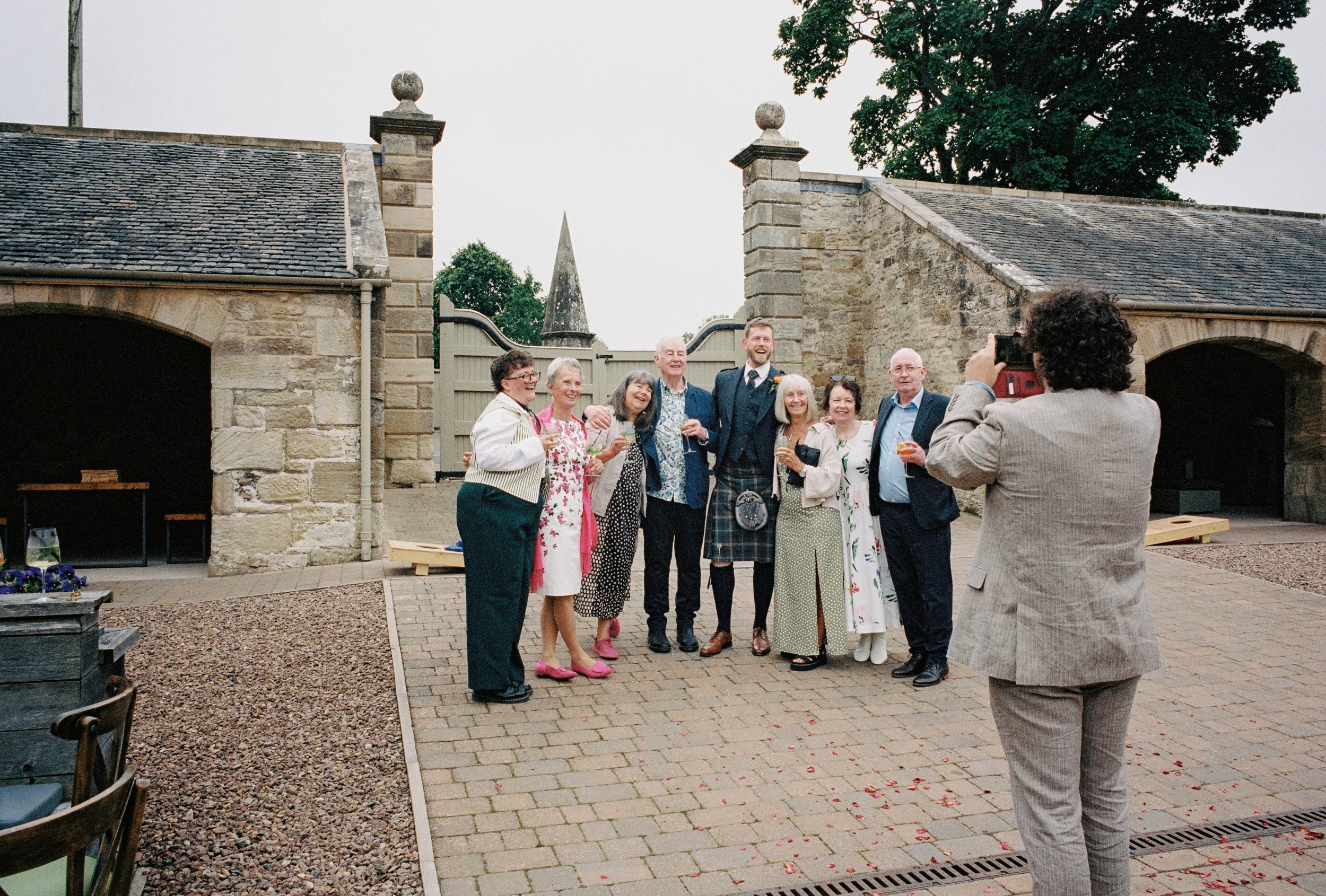 Group of people dressed in formal and semi-formal attire standing together outside a stone building, posing for a photo. One person in the foreground with curly hair and a gray plaid suit is taking their picture. The scene appears to be a celebration or special event, with some holding drinks and smiling.