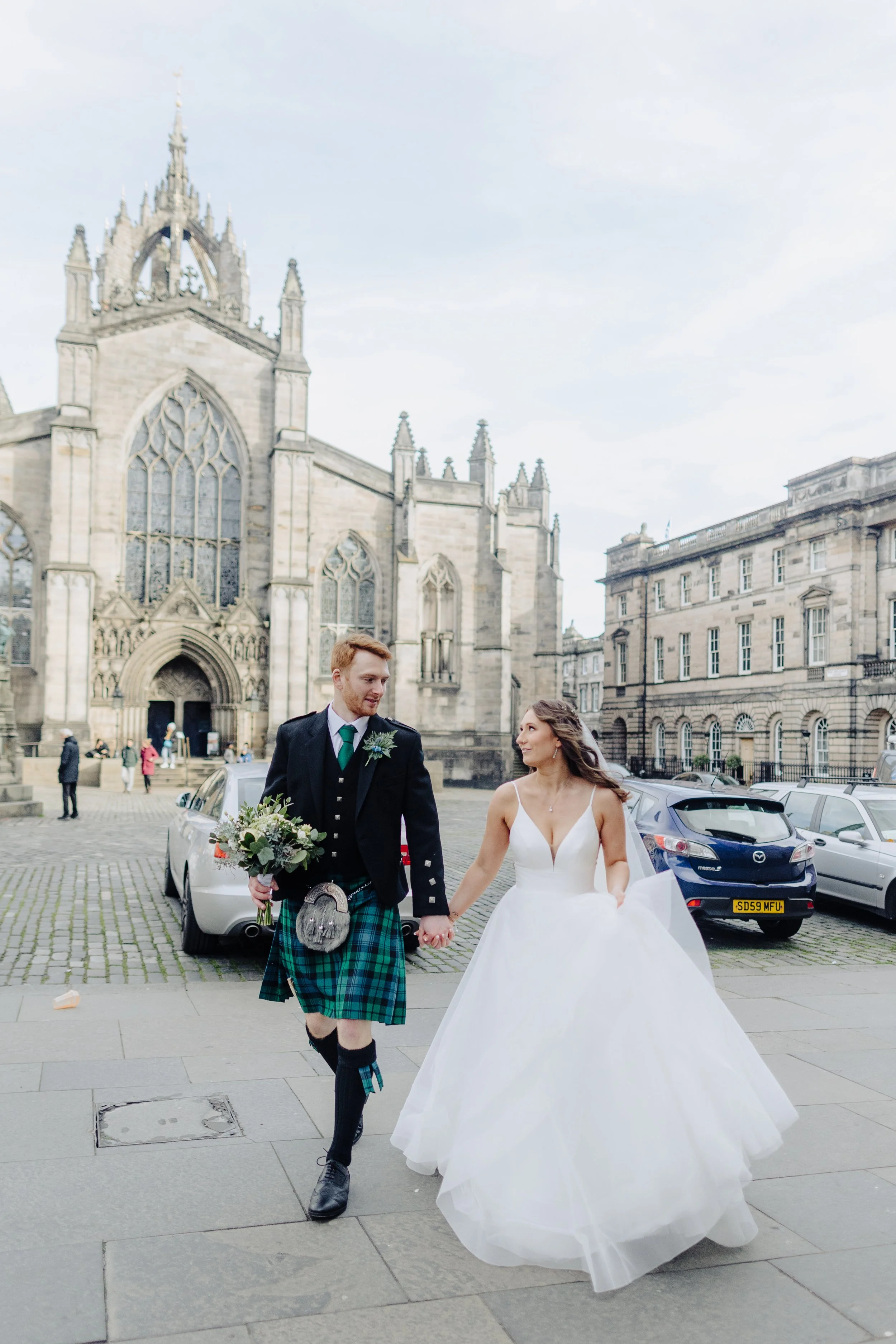 A newlywed couple walking hand in hand in front of a historic church, with cars parked along the street in an urban setting.