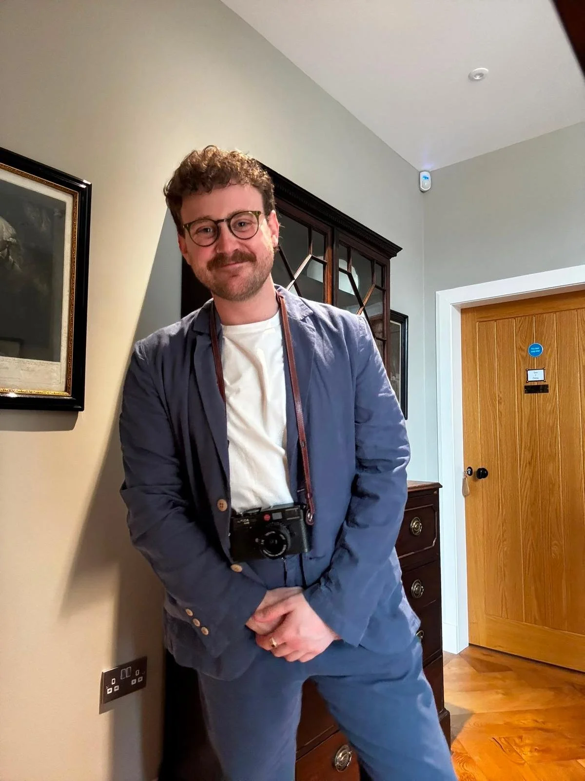 A man with curly hair, glasses, and a beard, smiling and wearing a blue blazer, white shirt, with a camera around his neck, standing in a room with wooden furniture and a closed wooden door.