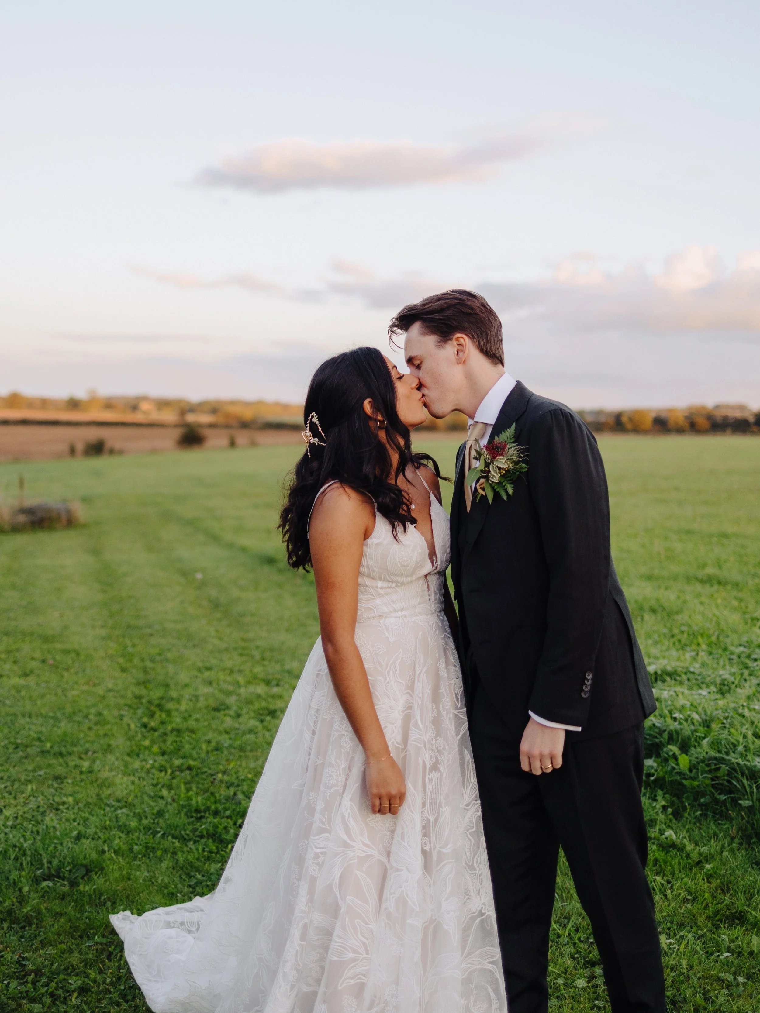 A bride and groom share a kiss outdoors on a grassy field during sunset. The bride wears a white lace wedding dress and has dark hair with a decorative hairpiece. The groom wears a dark suit, white shirt, and a boutonniere. The background features an open landscape with a partly cloudy sky.