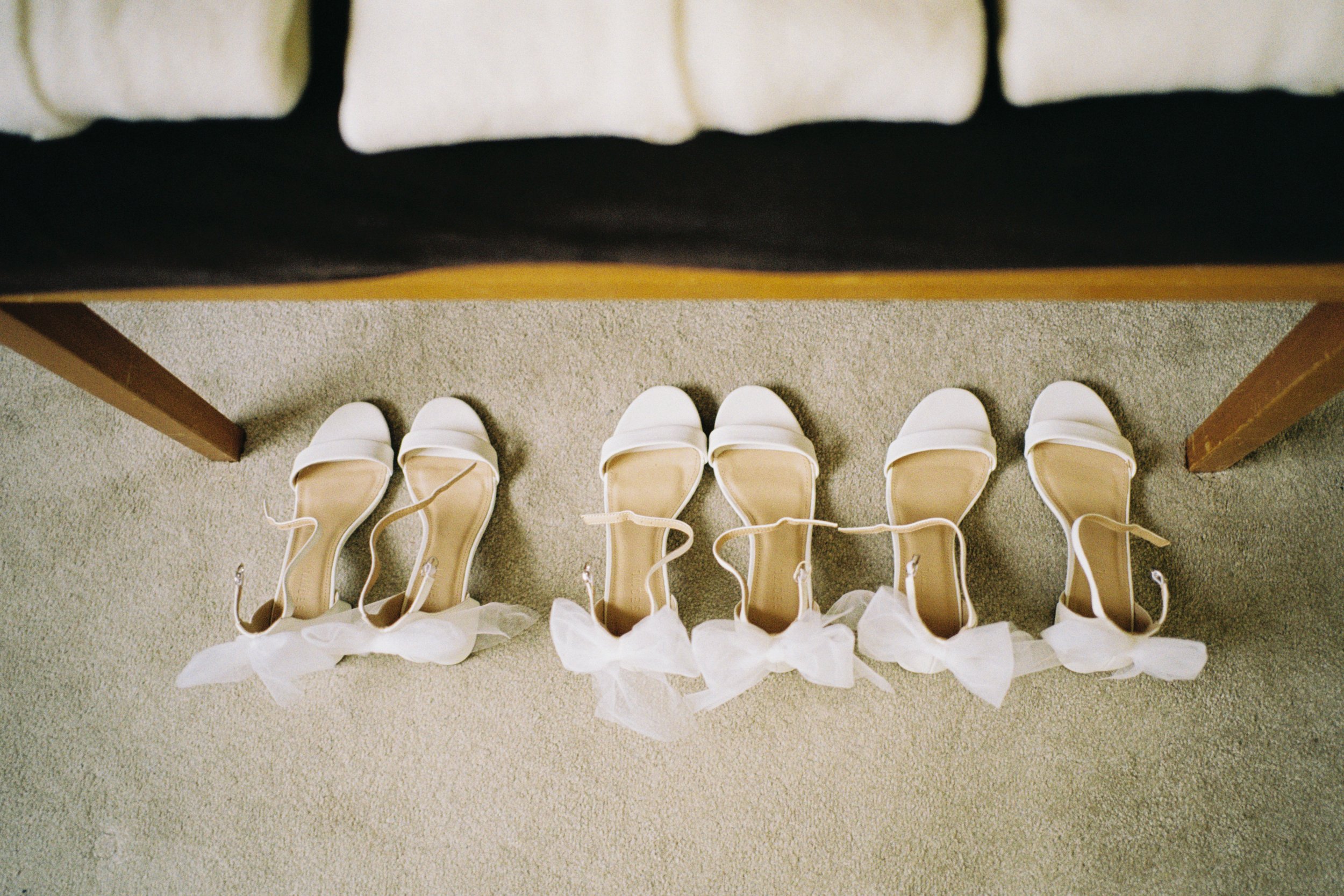 A top view of five pairs of white high-heeled shoes placed in a row on a beige carpet, with a person wearing a white wedding dress standing nearby.