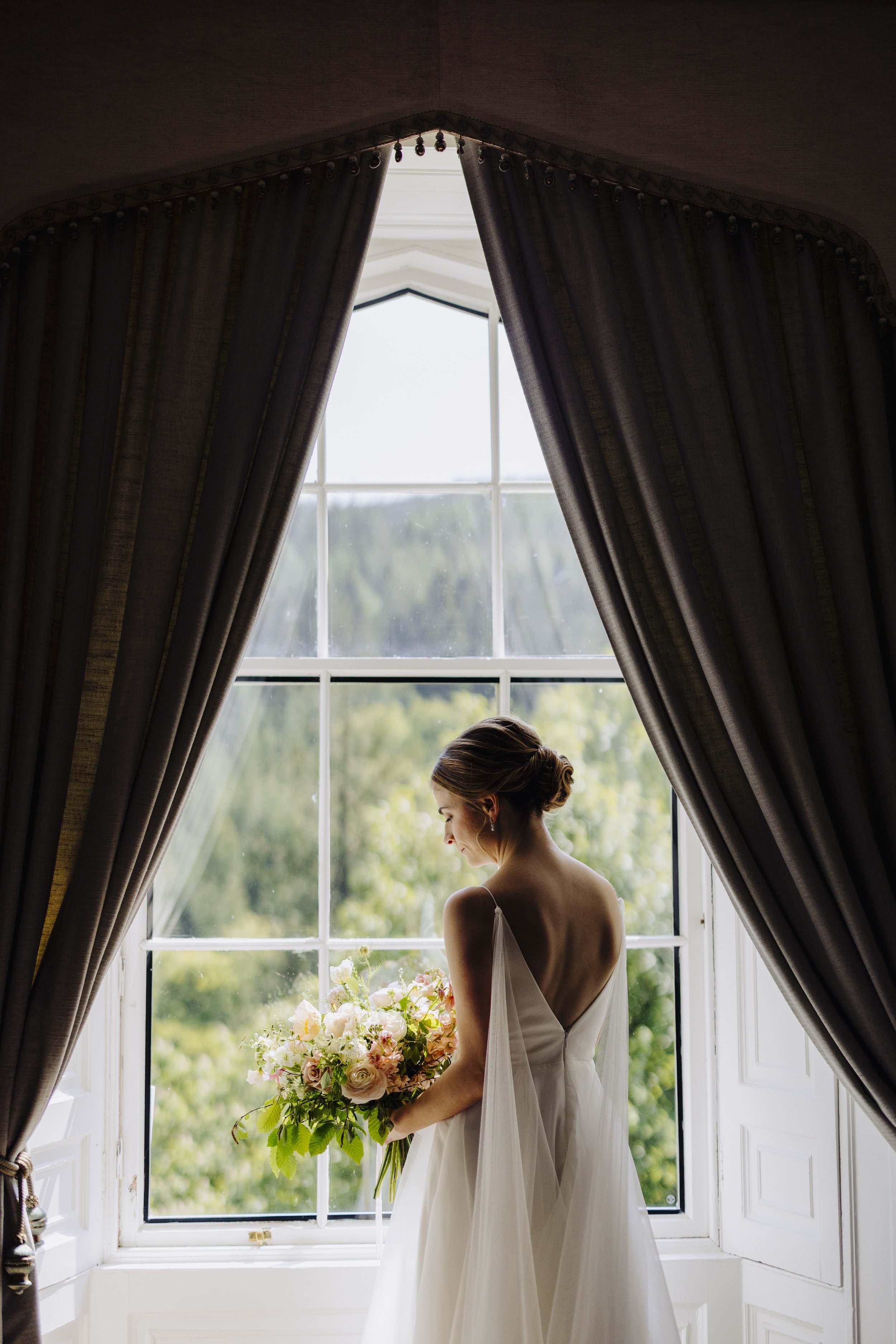 Bride with a strapless white wedding gown holding a large bouquet of pink and white flowers, standing by a window with curtains, natural light illuminating her face and dress.