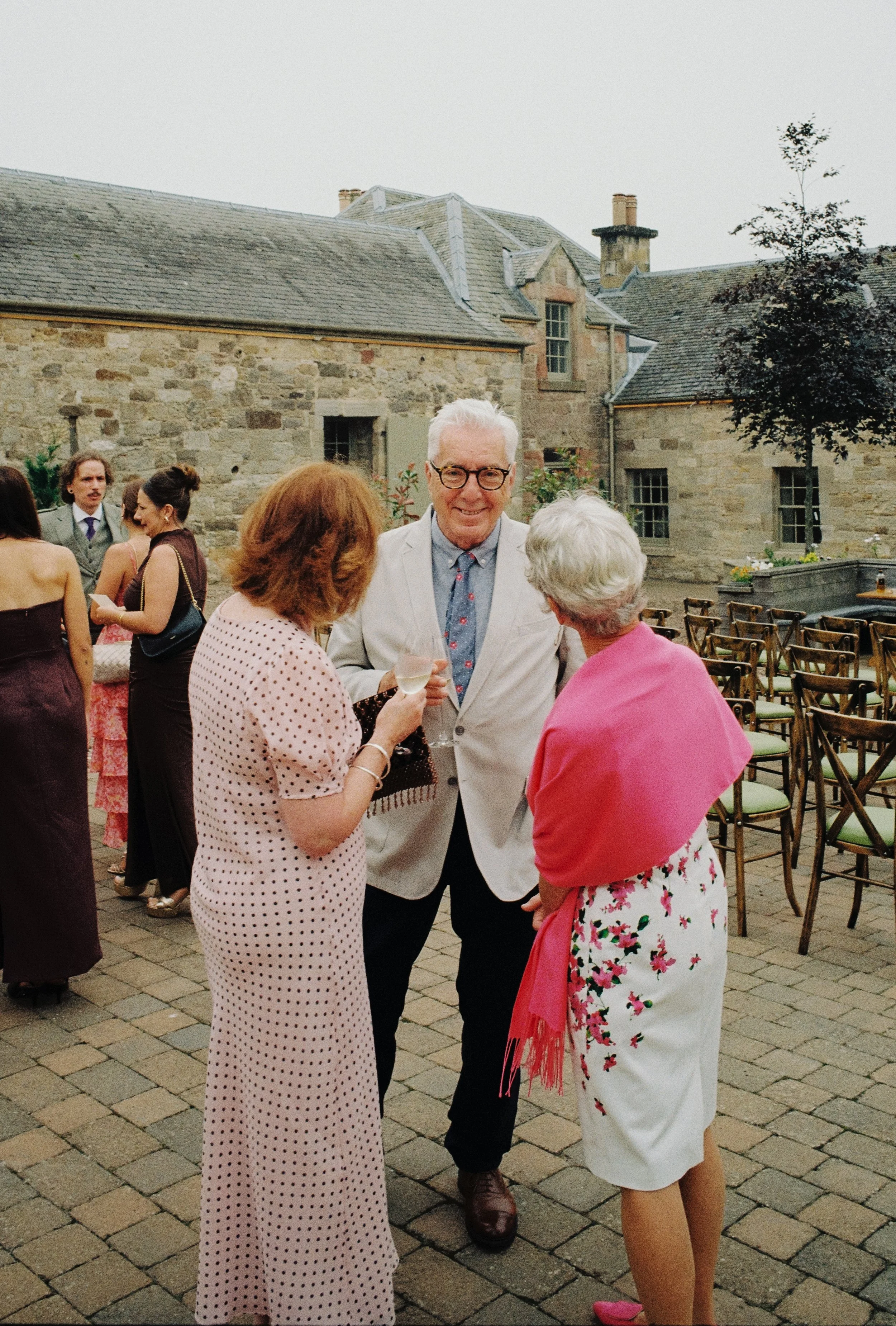 People gathered outdoors at a social event, standing and talking, with rows of chairs and a historic stone building in the background.