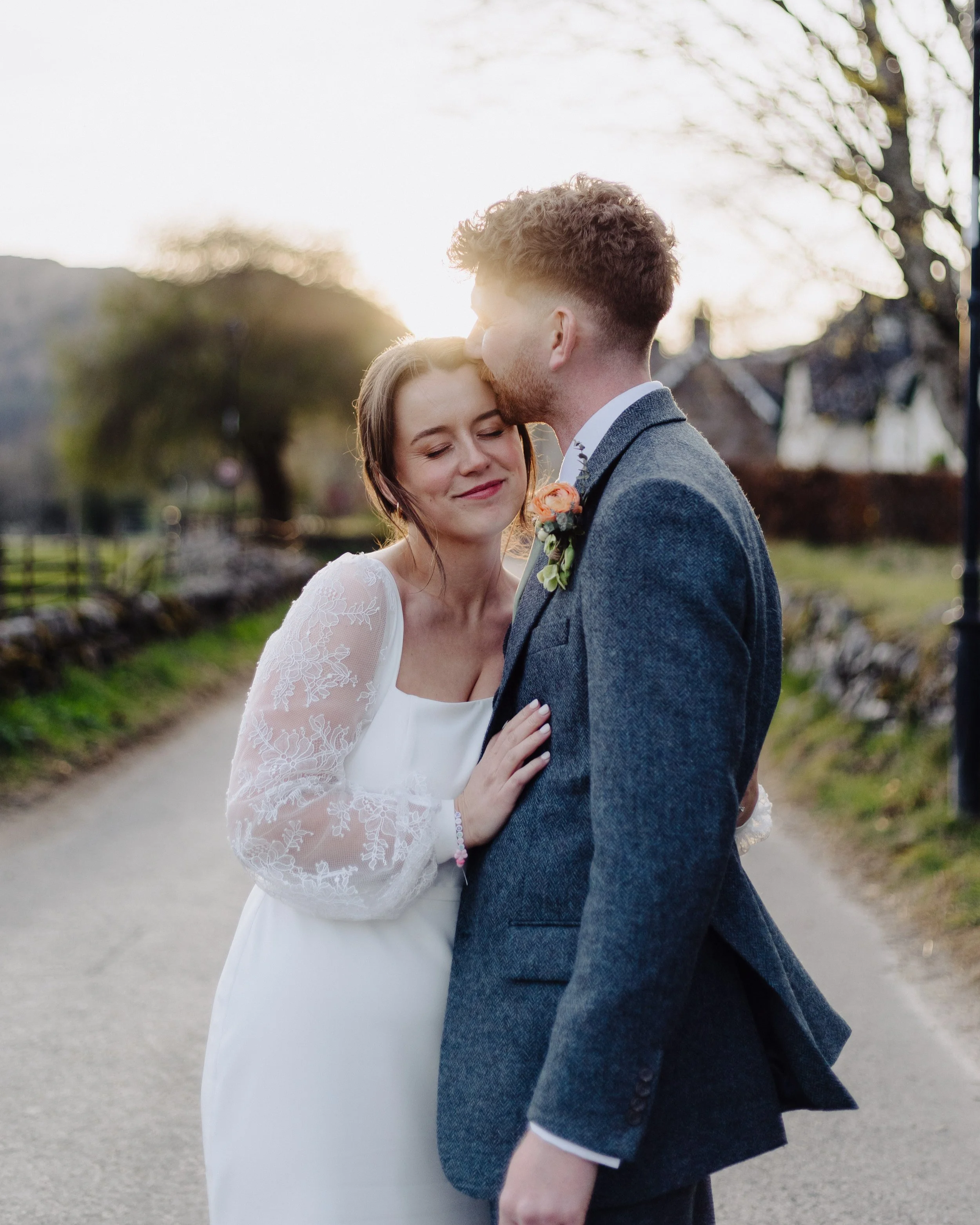 A bride and groom sharing a tender moment outdoors during sunset. The bride is smiling with her eyes closed, wearing a white lace wedding dress. The groom is kissing her on the forehead, dressed in a gray suit with a boutonniere. The background features a peaceful landscape with trees and a pathway.