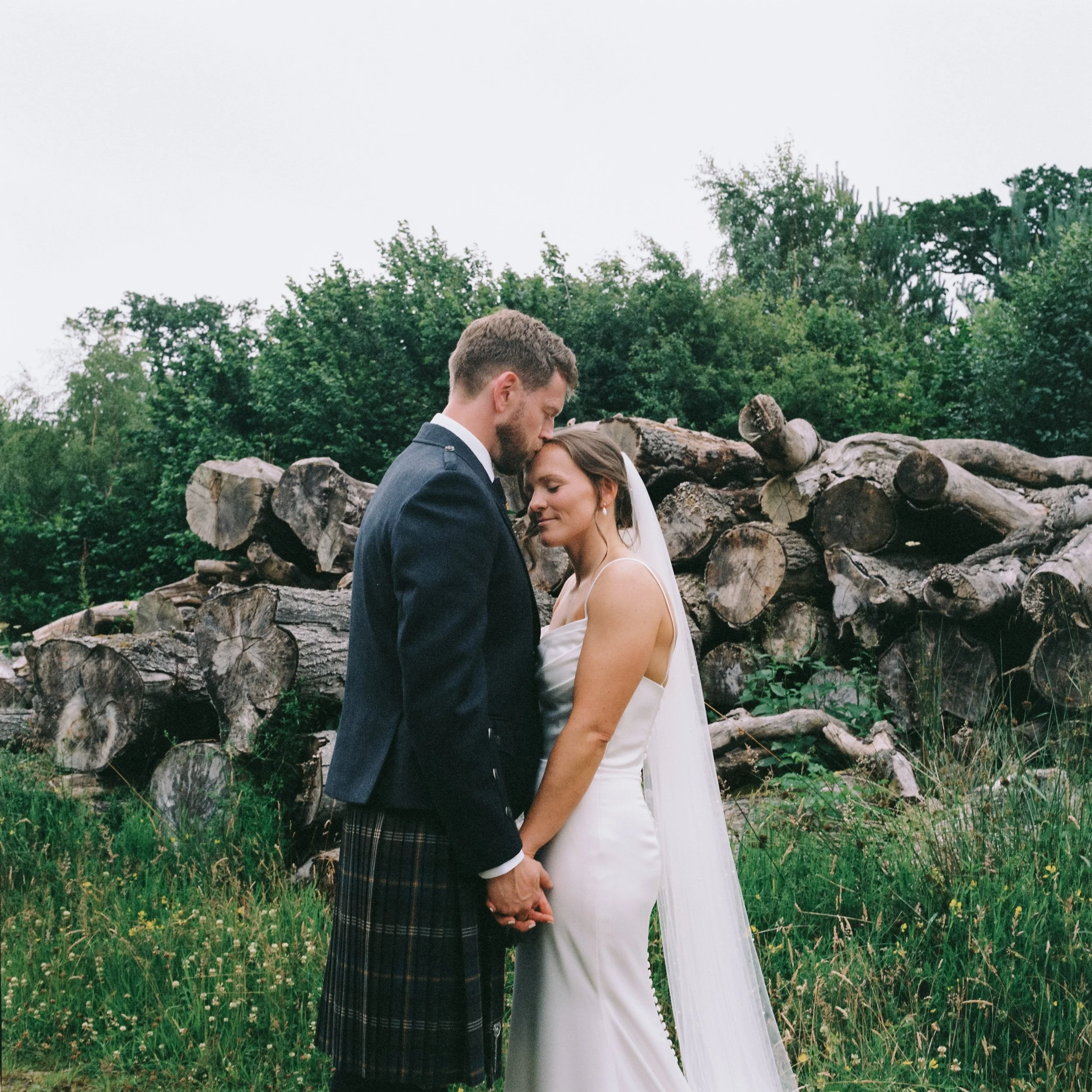 A man and woman in formal wedding attire kiss outdoors in front of a pile of logs with green trees and grass behind them.