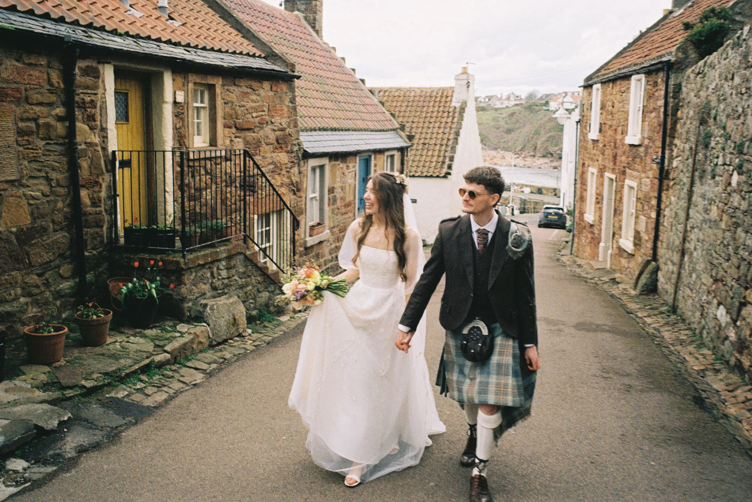 A bride and groom holding hands while walking down a street in a village with stone houses, some with flower pots and plants outside, on a cloudy day.