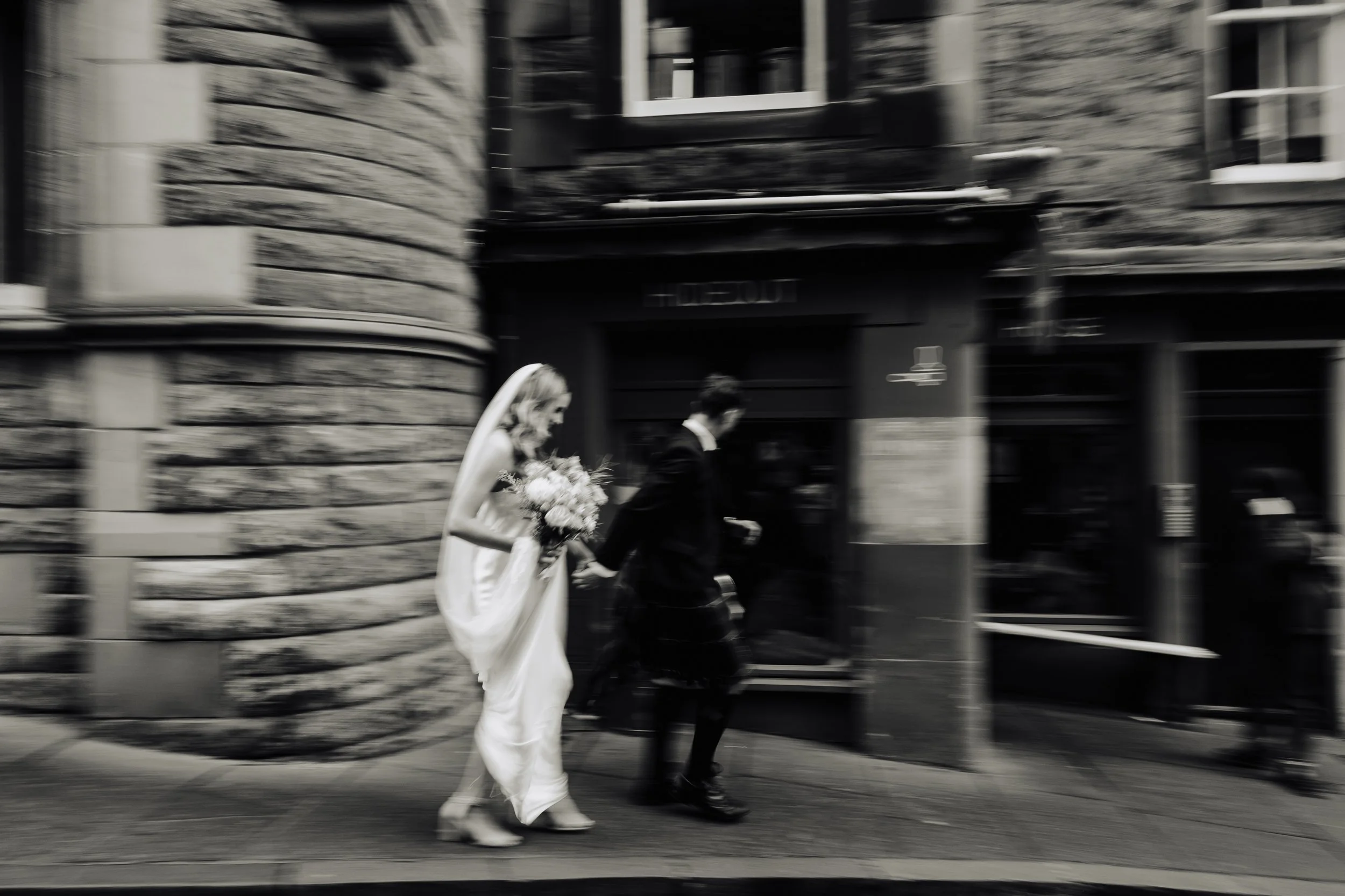 A black and white photo of a bride and groom walking hand in hand in front of a brick building, with motion blur. Royal mile Edinburgh