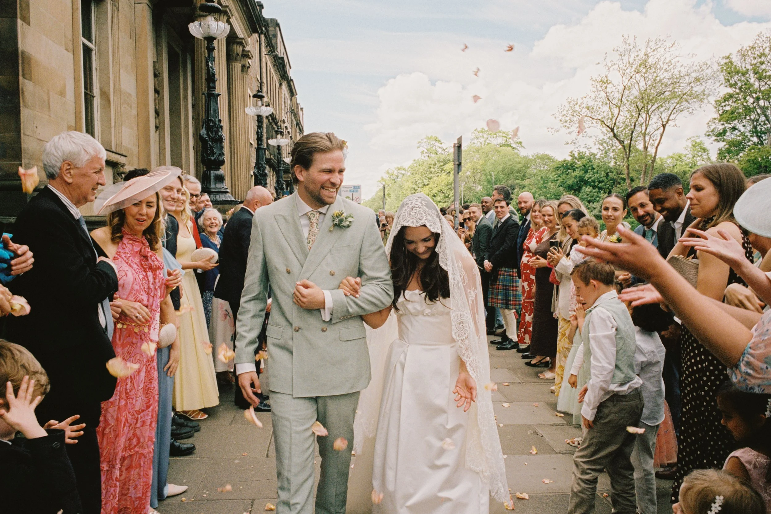 A bride and groom walking hand in hand through a crowd of wedding guests outside on a city street, with trees and historic buildings in the background, celebrating their wedding day.