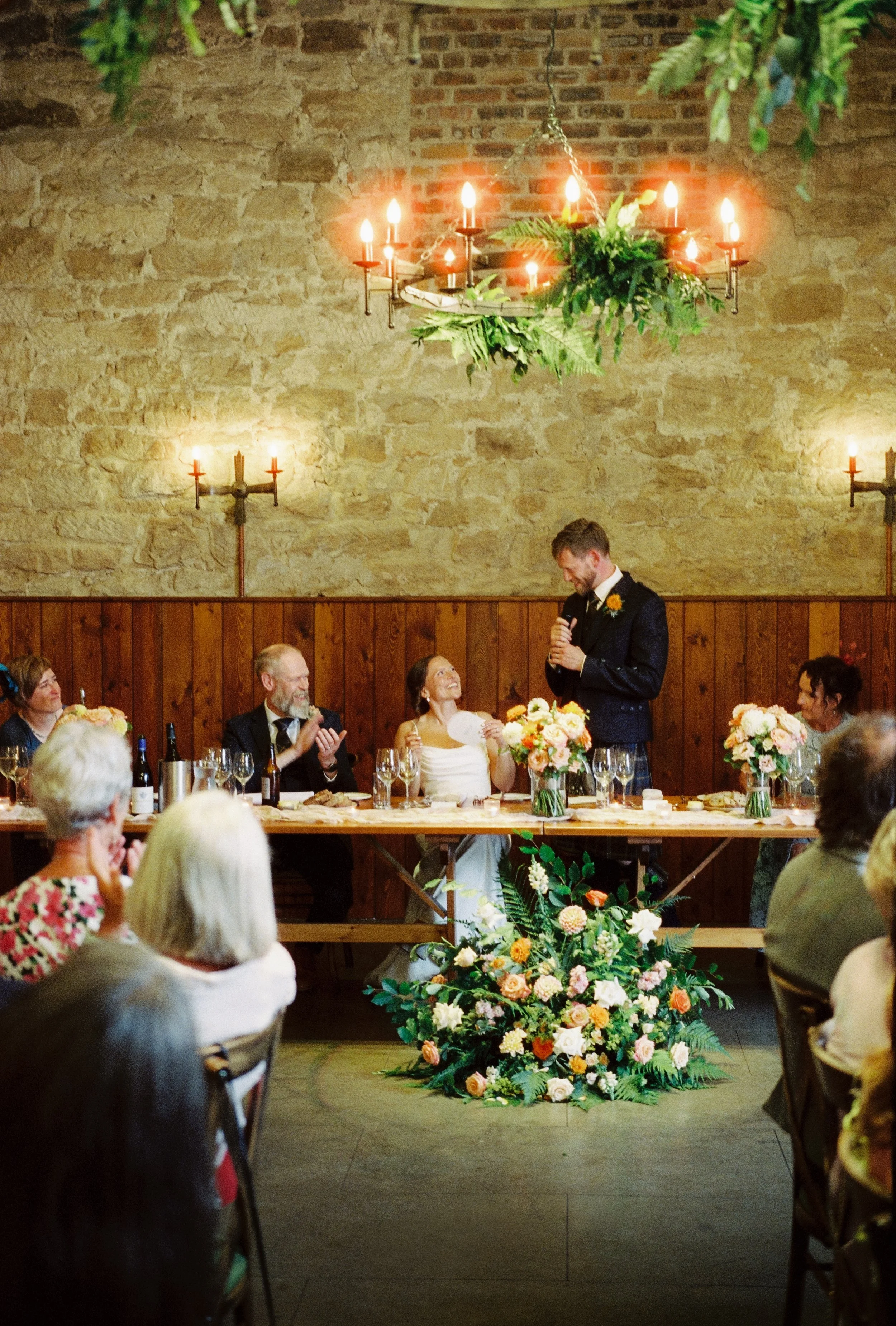 A wedding reception with a bride and groom sitting at the head table, surrounded by family and friends. A man in a tuxedo is giving a speech, with a bouquet of flowers in front. The setting has a rustic brick wall and a chandelier decorated with greenery and candles.