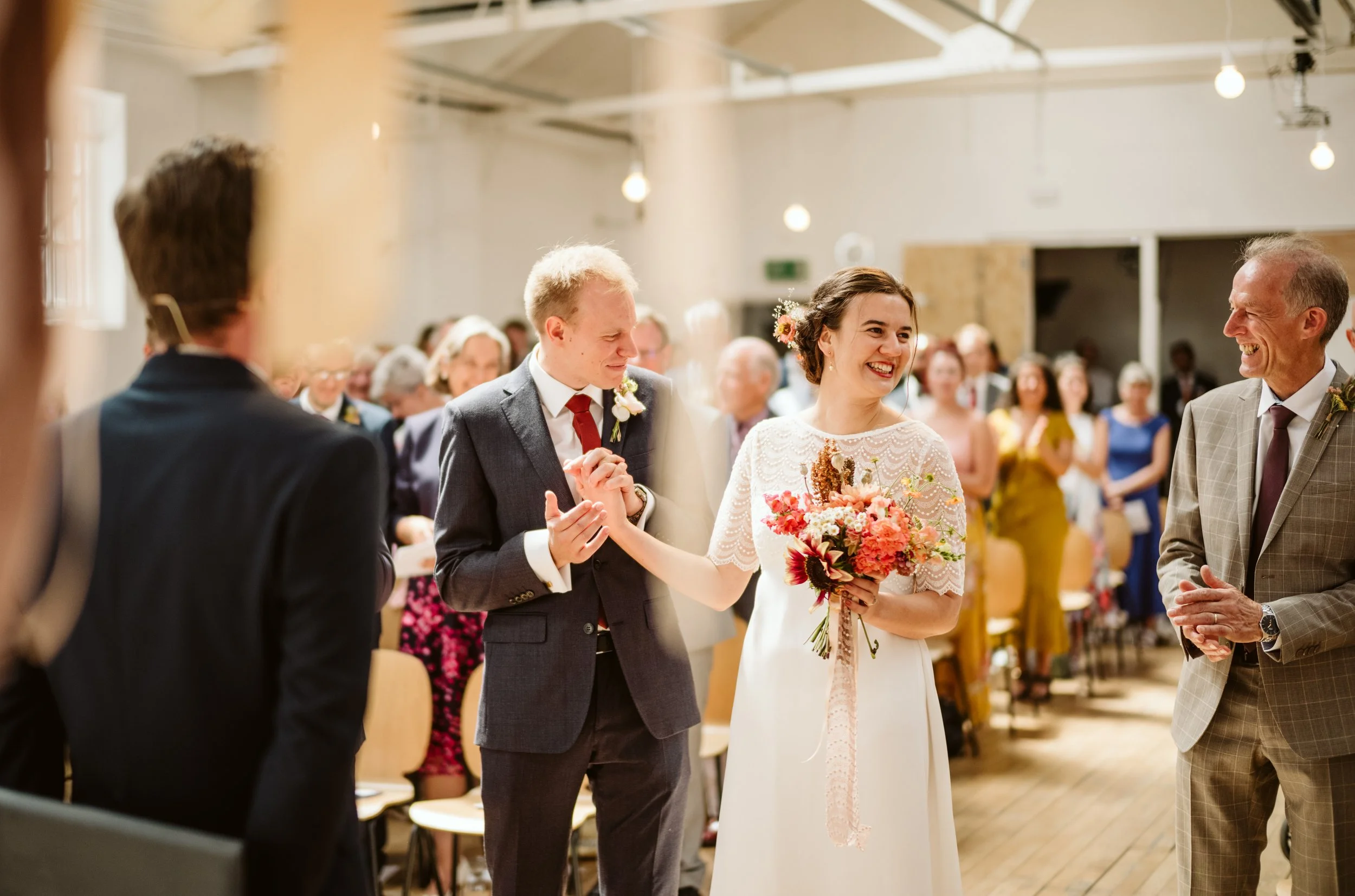 A wedding ceremony with a bride and groom holding hands, surrounded by family and friends. The bride is in a white lace dress holding a bouquet of flowers, and the groom is in a gray suit. They are smiling and appear happy, with guests seated and sta