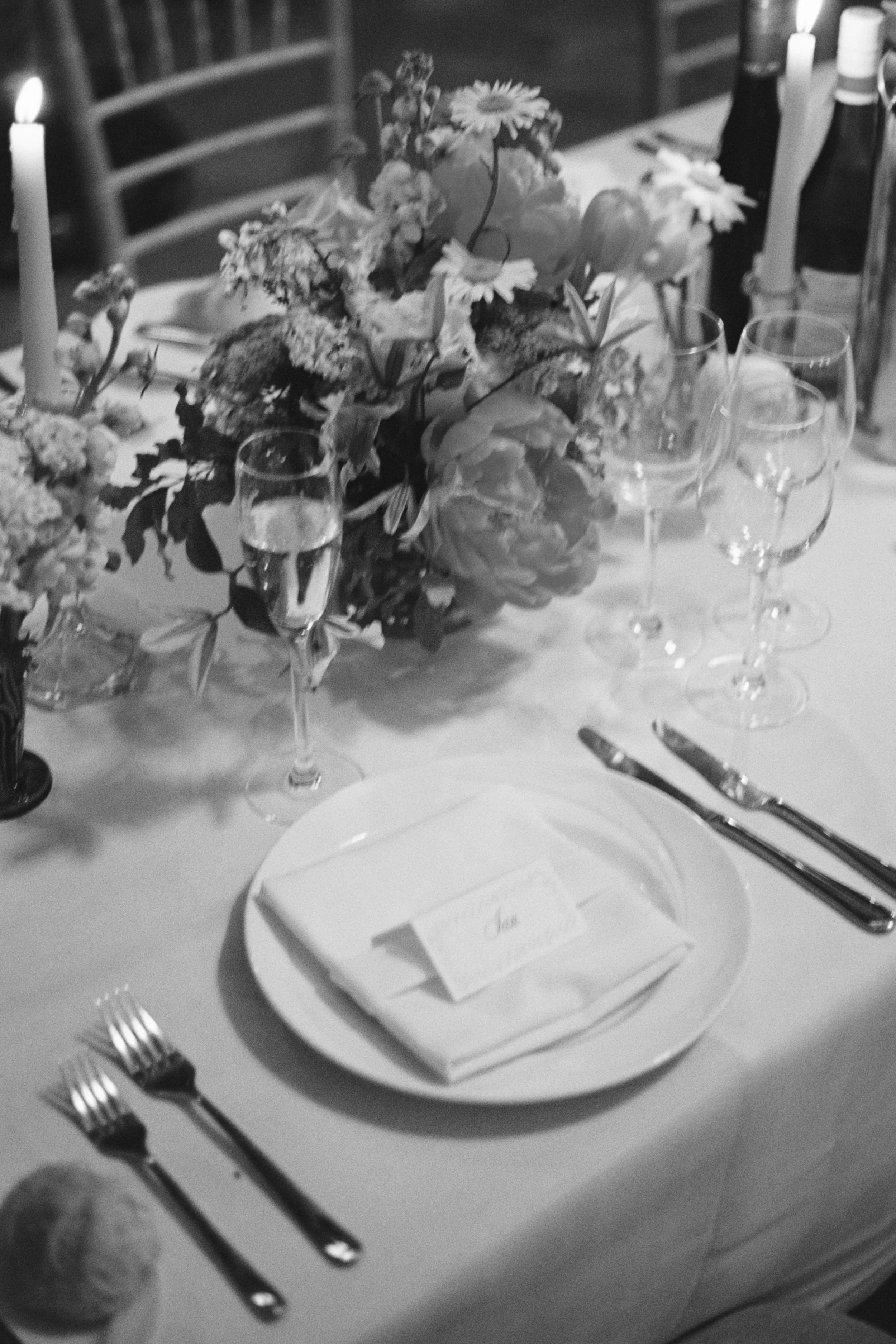 A dining table set for a formal event with a floral centerpiece, wine glasses, and elegant tableware. There are lit candles and a place card on the plate.