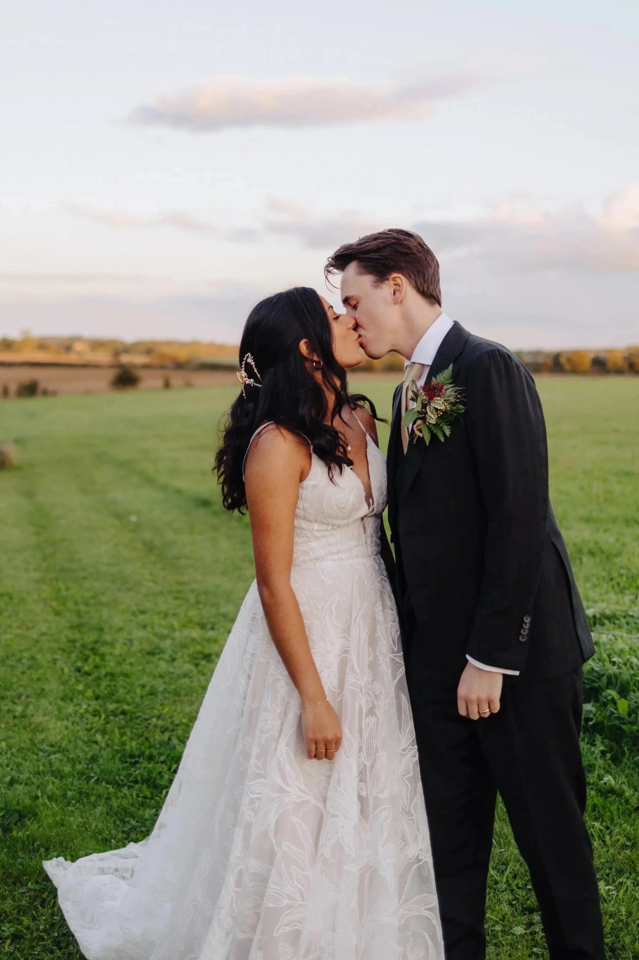 A bride and groom kissing outdoors on a grassy field during sunset, with a sky of light clouds in the background.