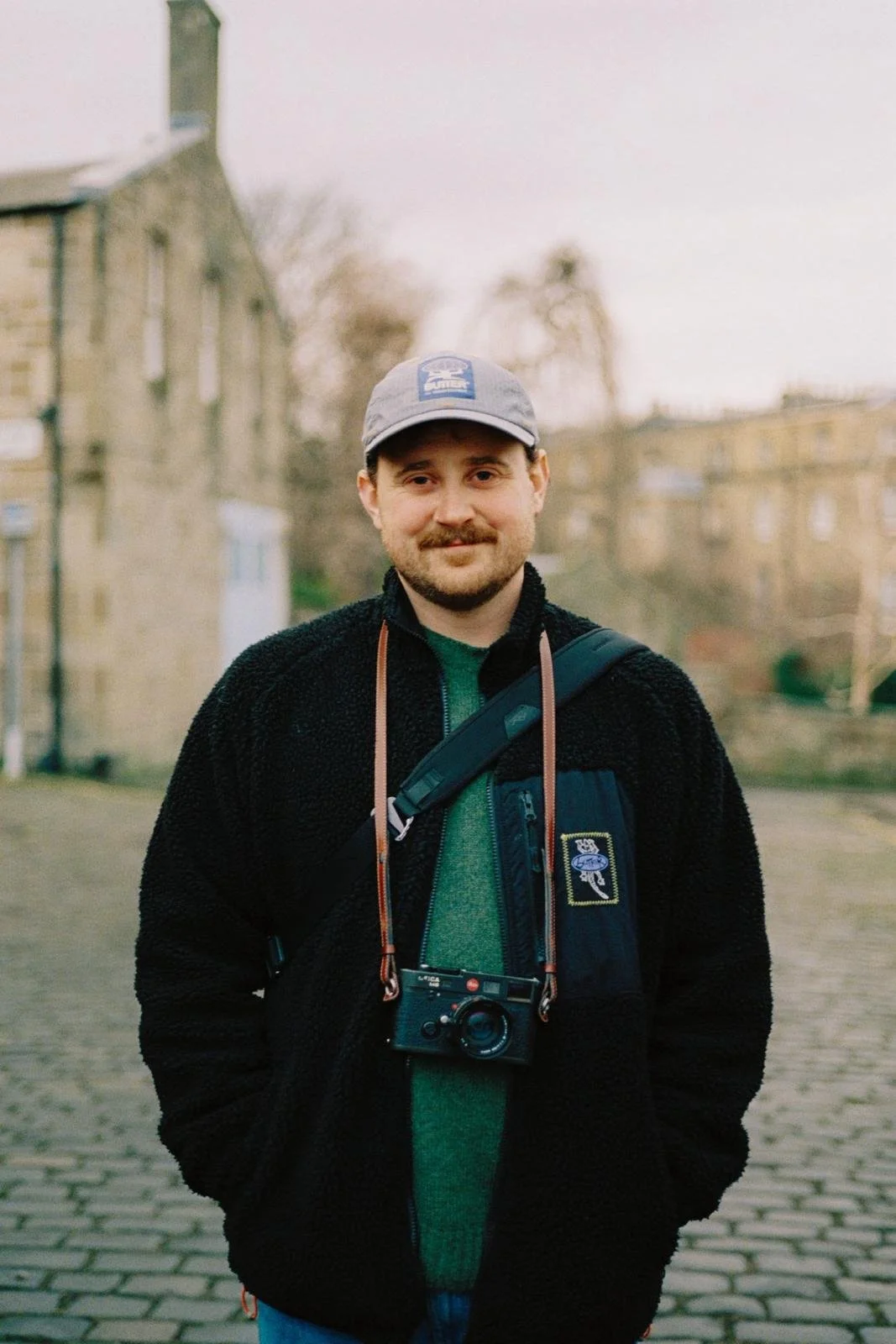 A man with a beard and mustache, wearing a gray cap, a green shirt, and a black fleece jacket, standing outdoors on a cobblestone street with buildings and trees in the background, carrying a camera around his neck and a black crossbody bag.