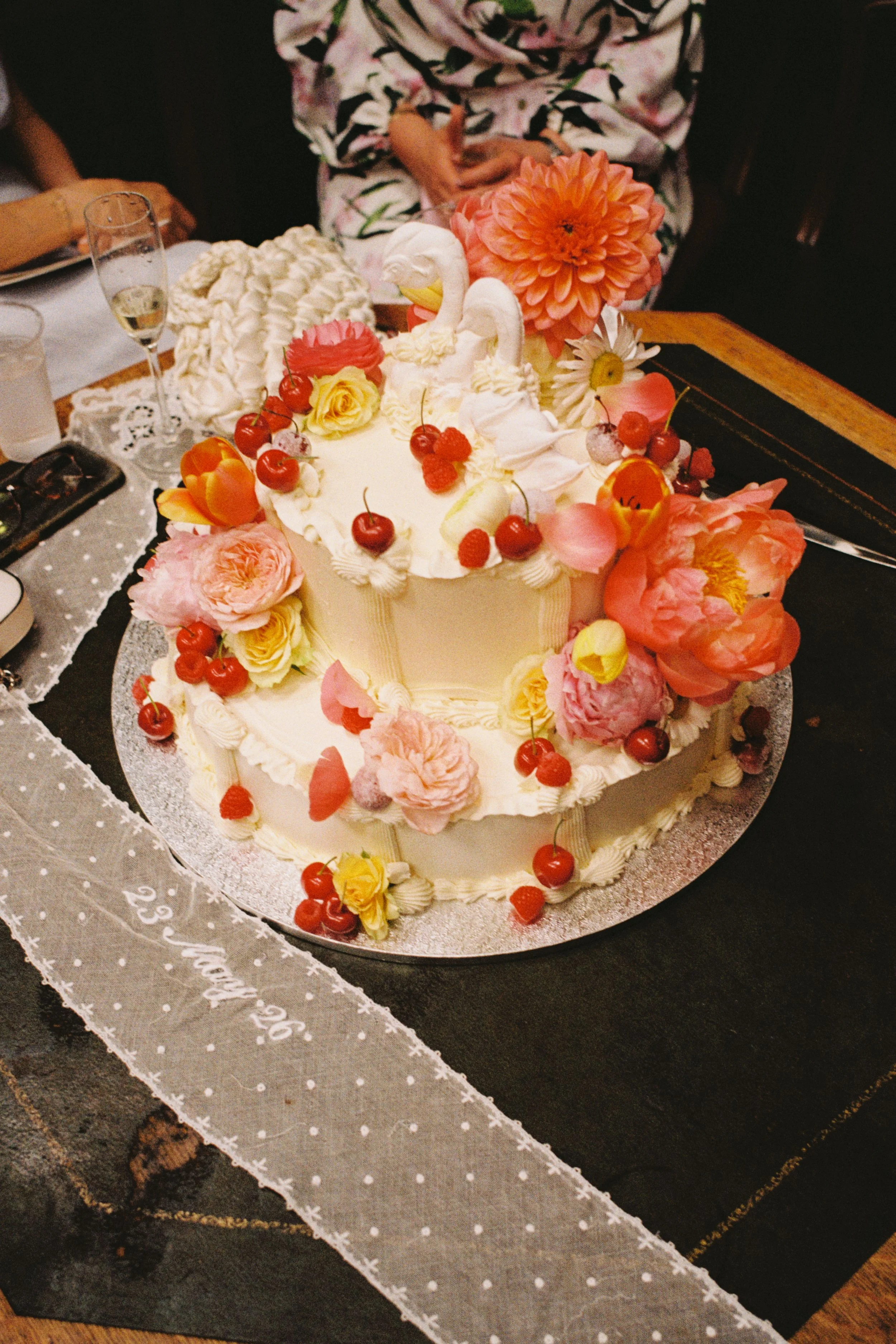 A decorated birthday cake with white frosting, topped with colorful flowers, cherries, and swan-shaped decorations, surrounded by people celebrating at a table.