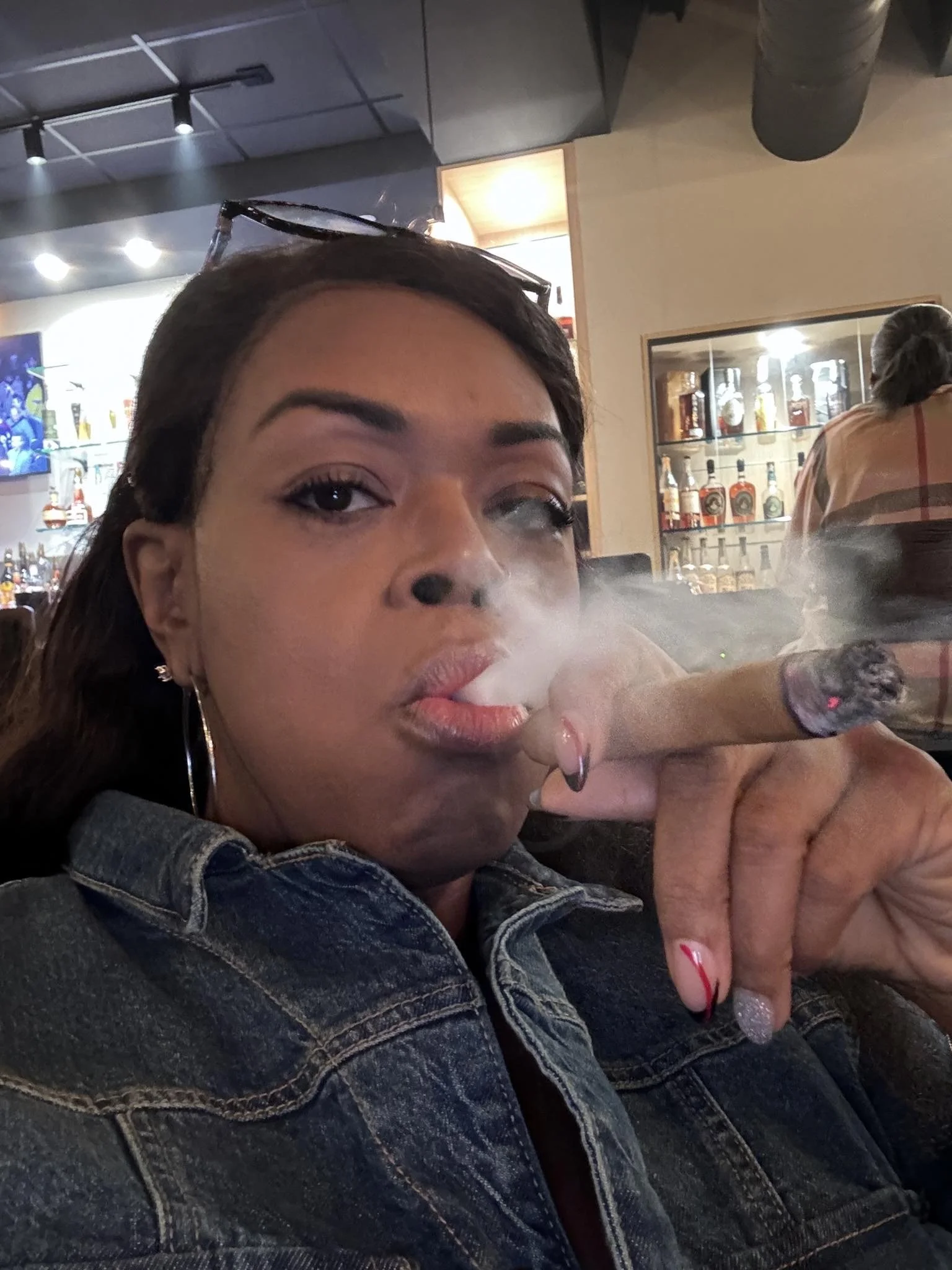 Black woman with glasses sitting on top of her head blows out smoke with a cigar in her hand