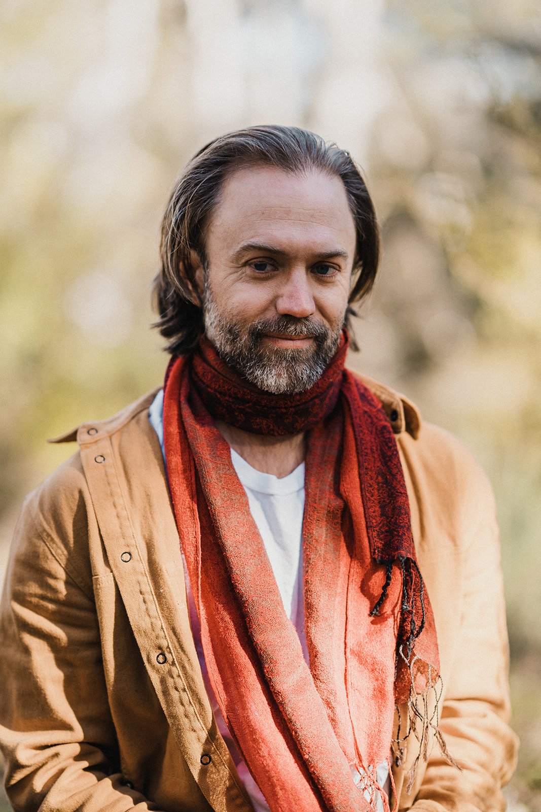 Portrait of a man wearing a red scarf and tan jacket outdoors in nature with a calm expression and blurred forest background.