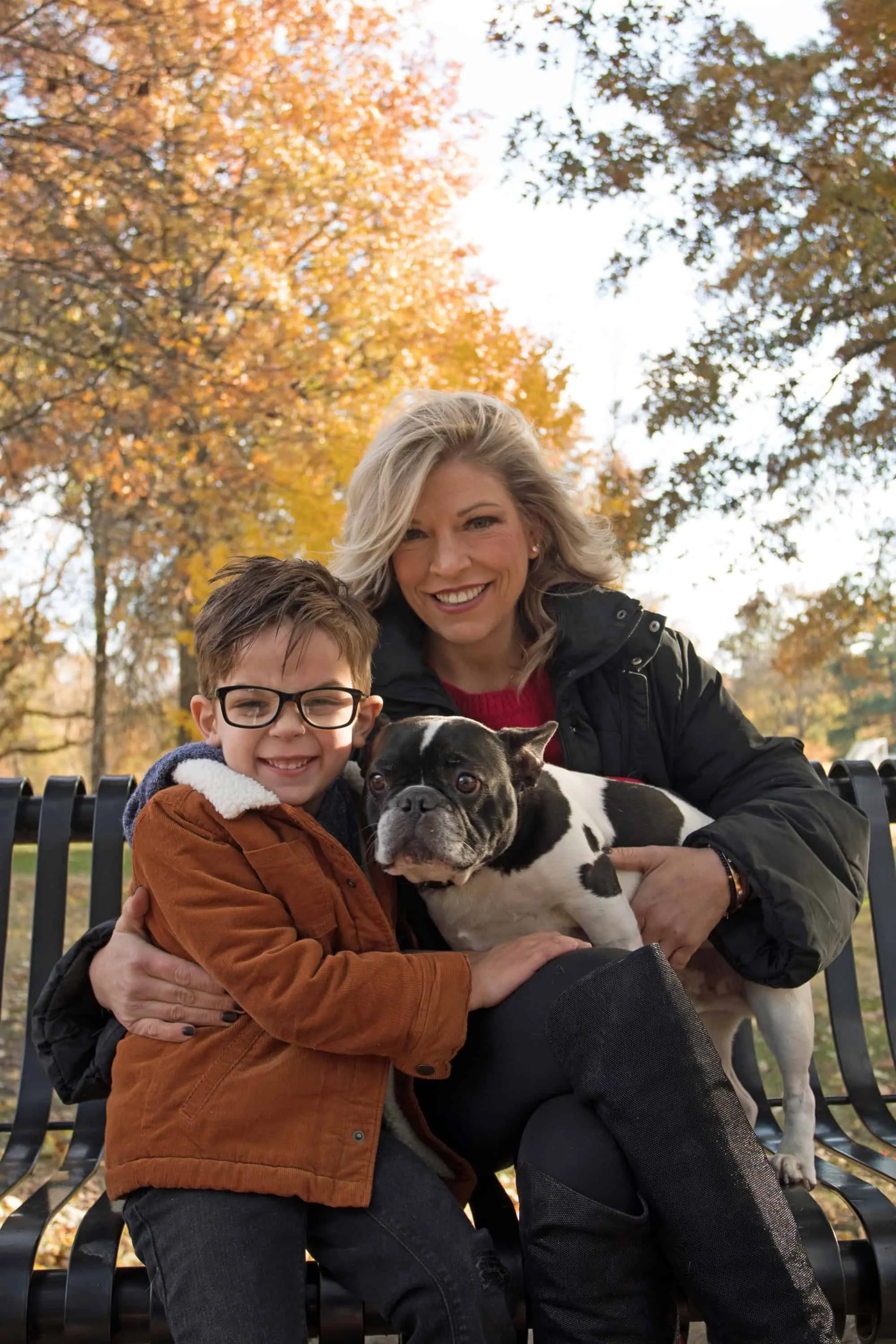 Smiling mother stands outside with her young son and dog surrounded by colorful fall trees.