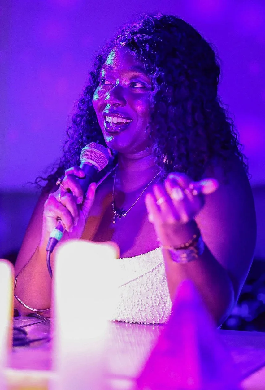 Delanyo Mensah speaks into a handheld microphone while gesturing with her other hand. She wears a white strapless top and is illuminated by purple and blue stage lighting, with a bright light in the foreground.