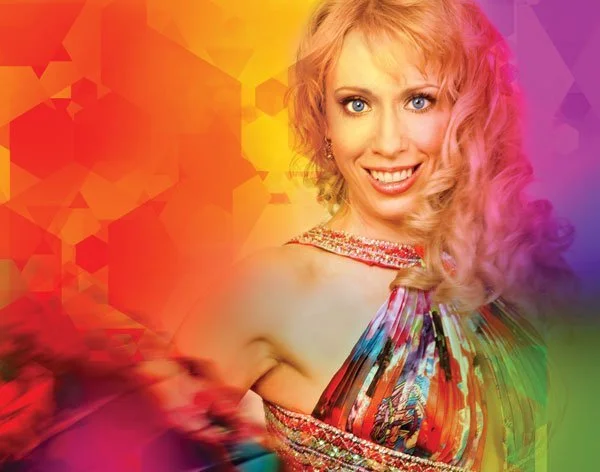 Portrait of a smiling blonde woman in a colorful dress against a vibrant rainbow background.