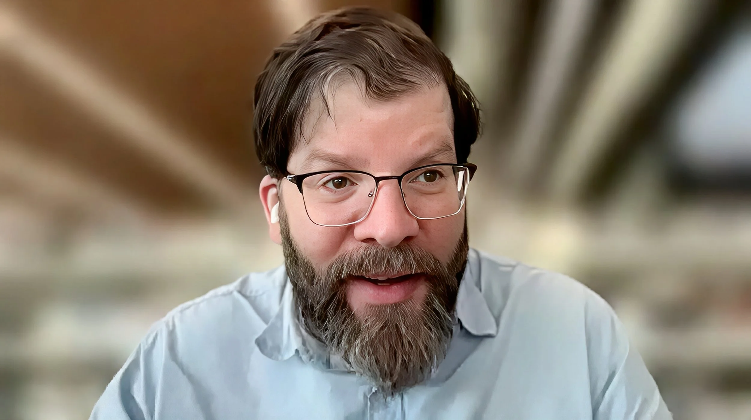 A bearded man wearing glasses, white wireless earbuds, and a light blue button-up shirt smiles toward the camera during a video interview, with a softly blurred indoor background.