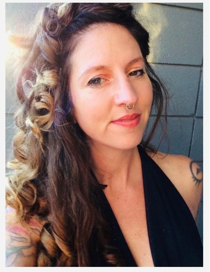 Close-up portrait of a woman with long wavy hair, nose ring, and tattoos outdoors.