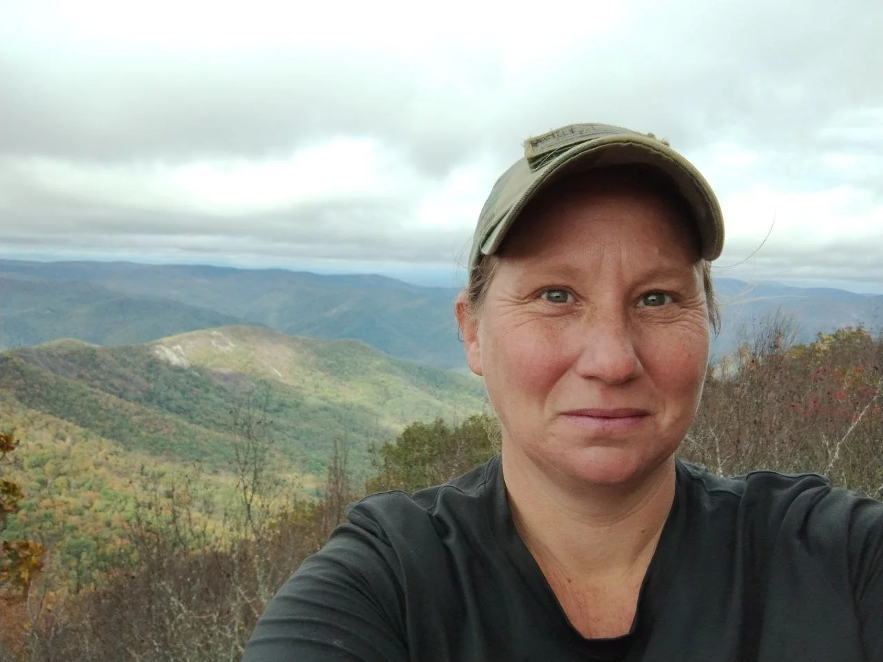 A woman wearing a baseball cap takes a selfie in front of a cloudy mountain overlook.