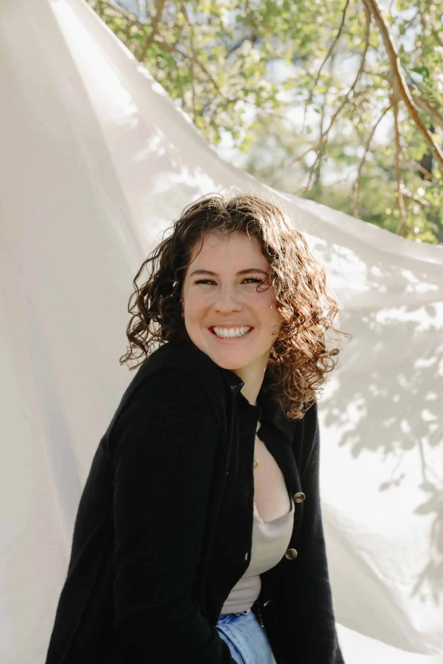 brunette woman with short curly hair sits in front of a white sheet outside and smiles at the camera