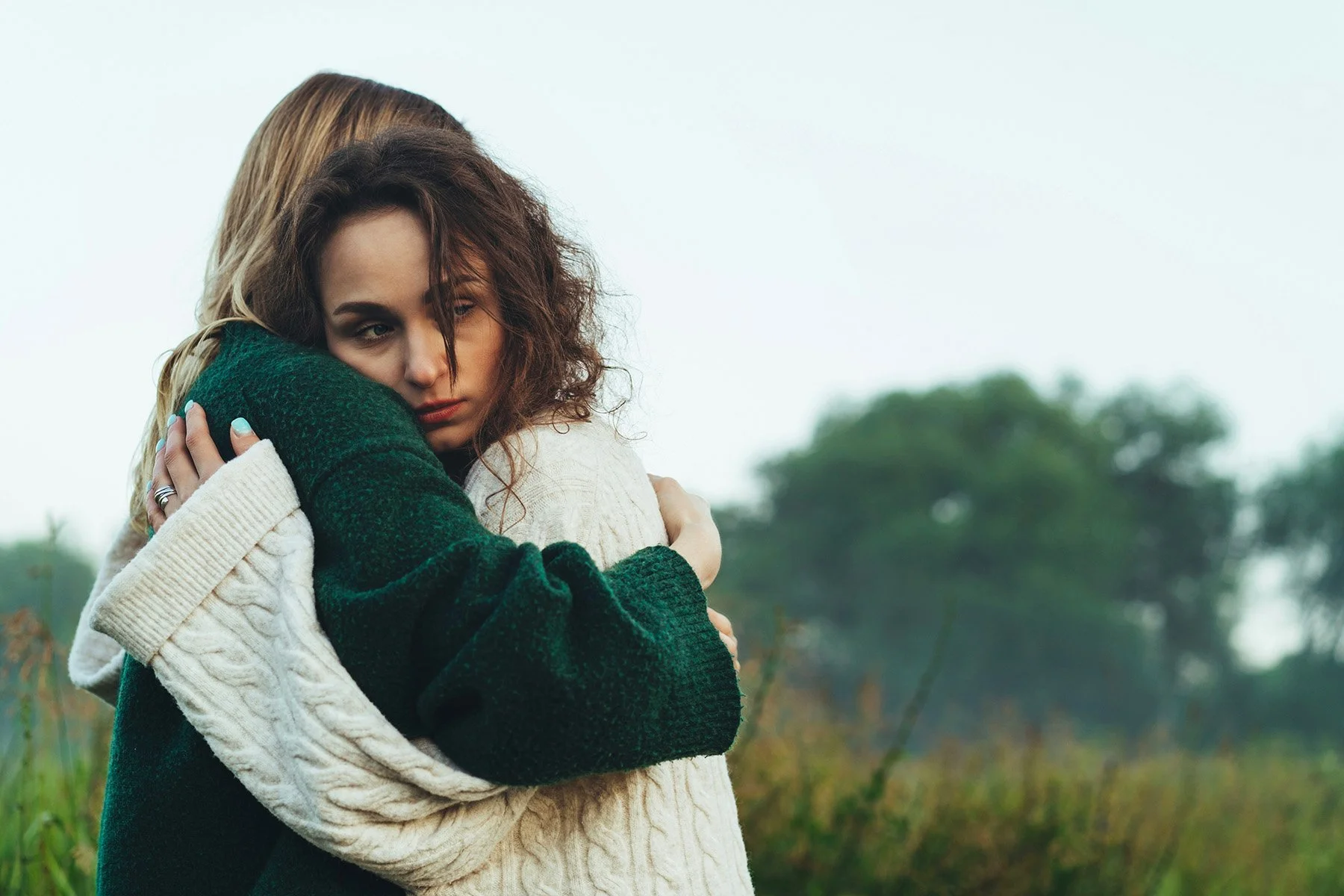 Two people wearing knit sweaters hug outdoors with trees and a grassy field in the background.