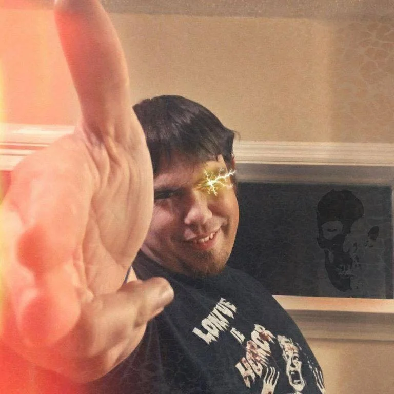 Man smiling while reaching a hand toward the camera with a playful visual effect over one eye, wearing a graphic T-shirt indoors.