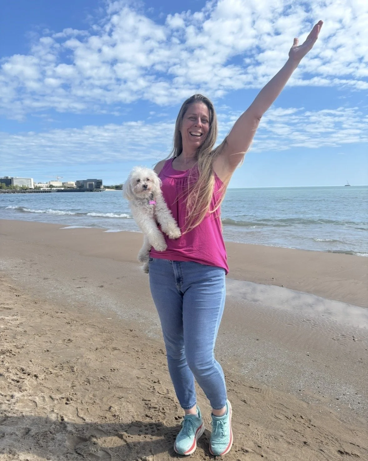 A woman stands on a sandy beach holding a small white dog in one arm and raising the other arm toward the sky, smiling, with water and a partly cloudy sky behind her.