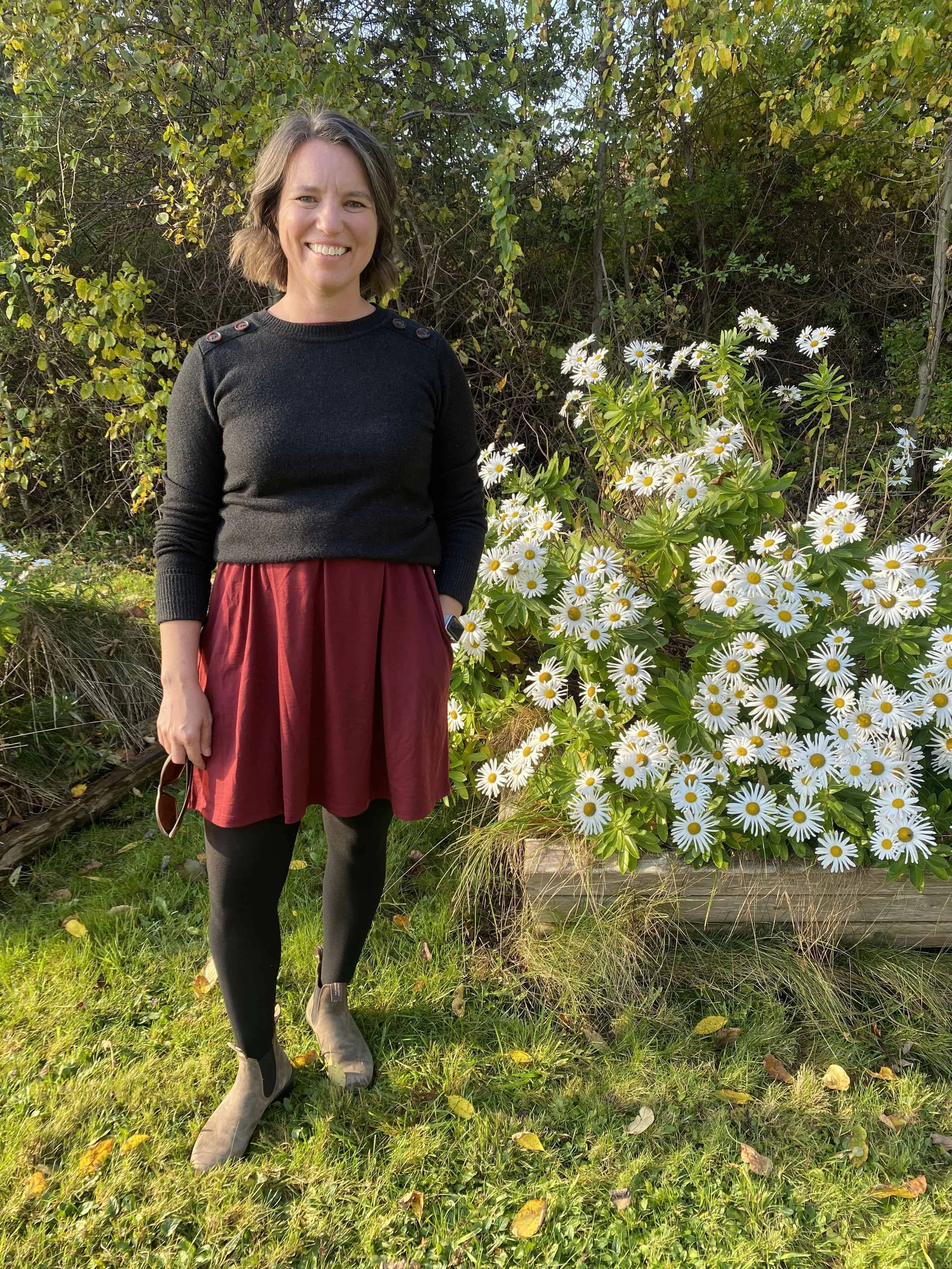 Brunette woman in a black top and maroon skirt smiling in front of a bed of daisies.