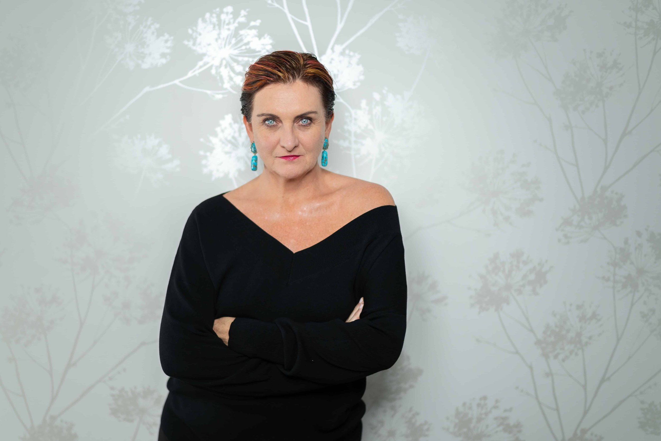 Simone Milasas standing with her arms crossed in a black off-the-shoulder top, looking directly at the camera against a pale botanical background.