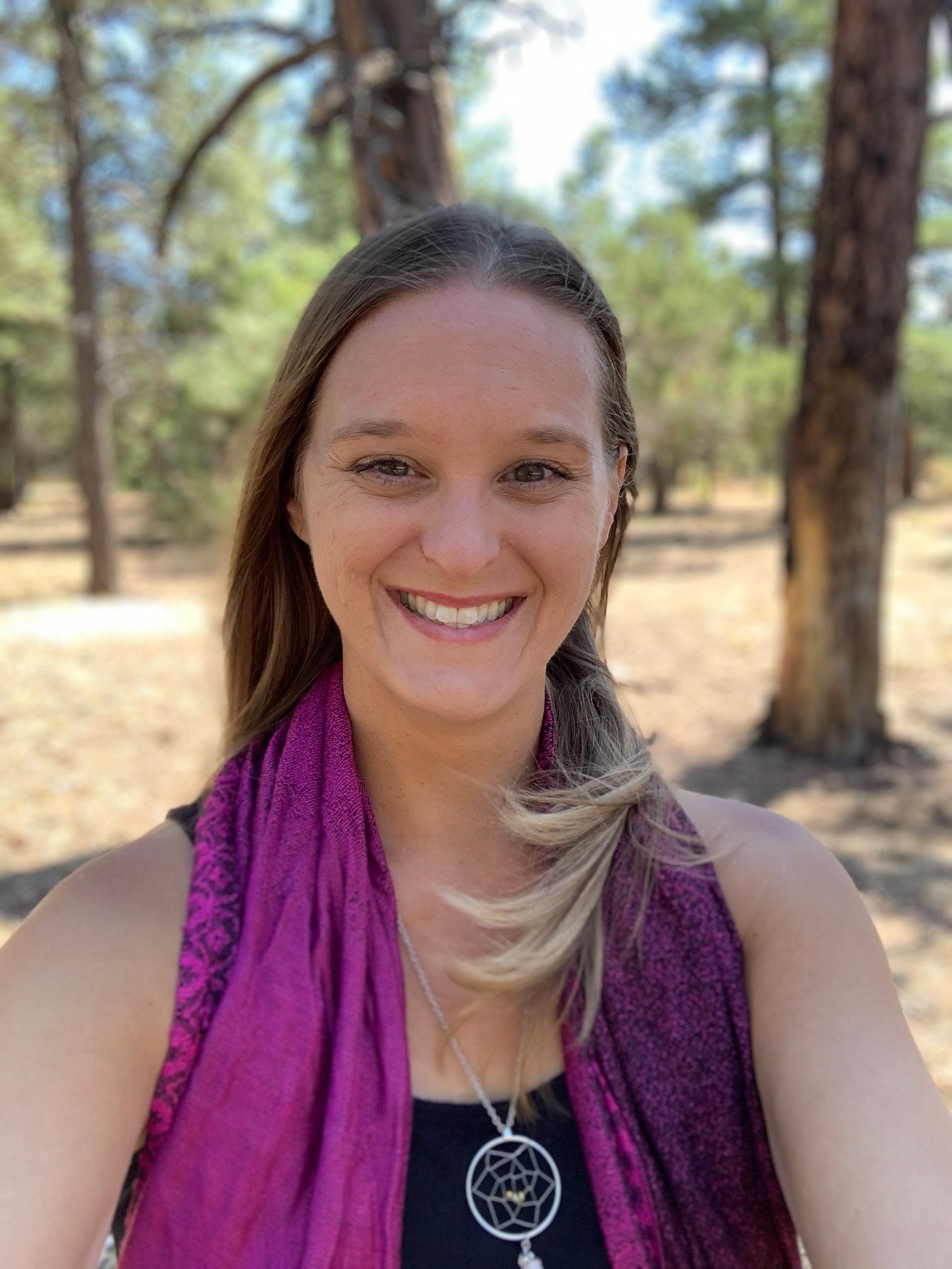 Smiling woman outdoors in a wooded area wearing a purple scarf and geometric pendant necklace.