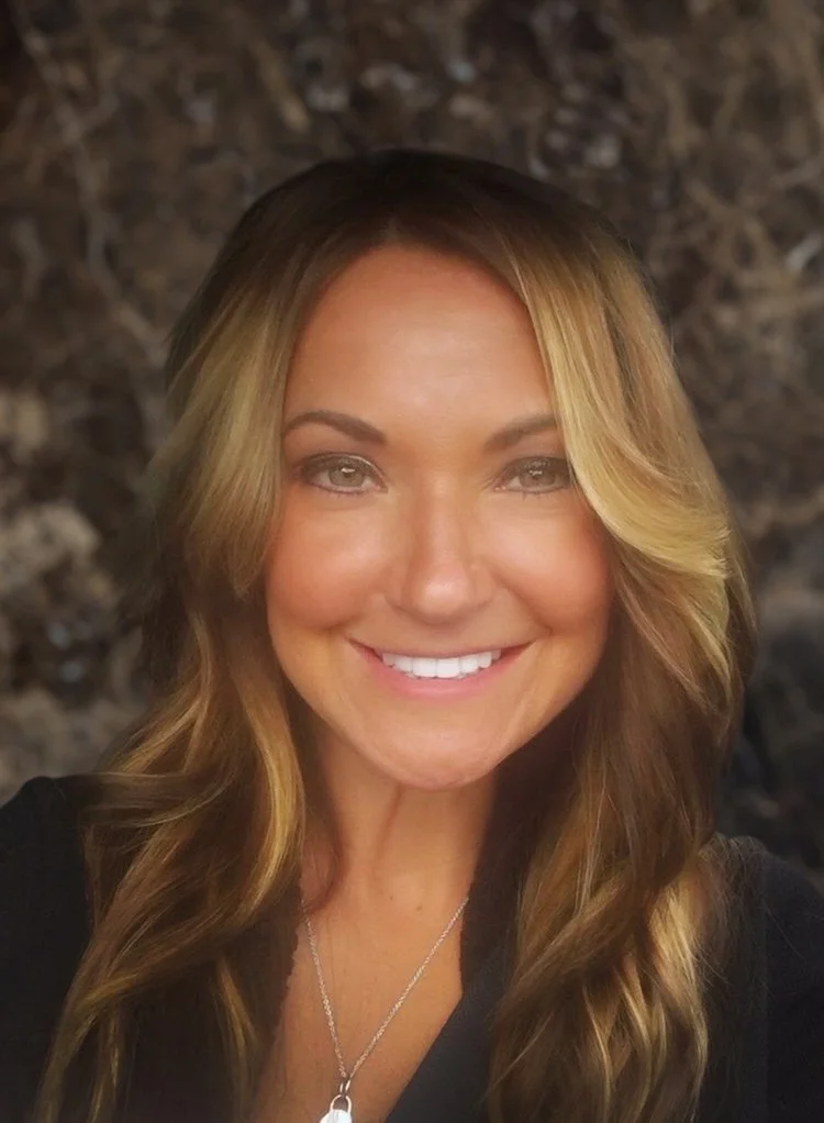 Smiling woman with long highlighted brown hair wearing a black top and silver necklace, standing in front of a textured rock wall.
