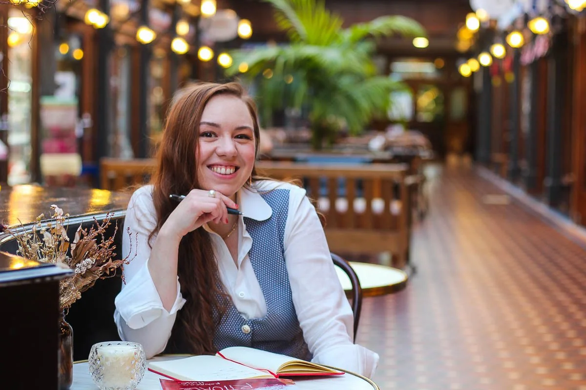 Happy woman sitting at a table with her chin resting on her hand while holding a pen beside an open notebook.
