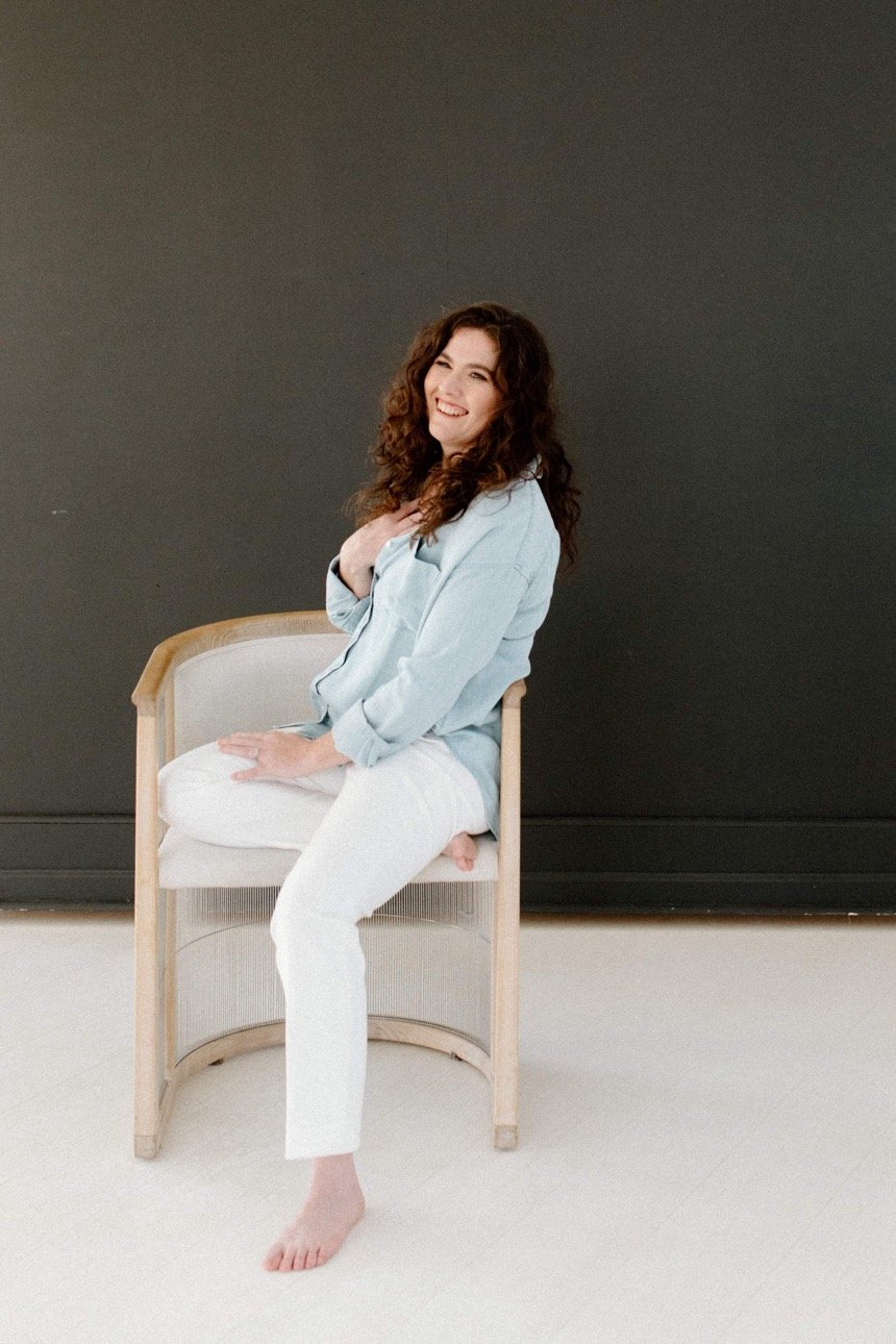 Curly haired brunette woman smiles, posing on a chair with her hand to her heart.