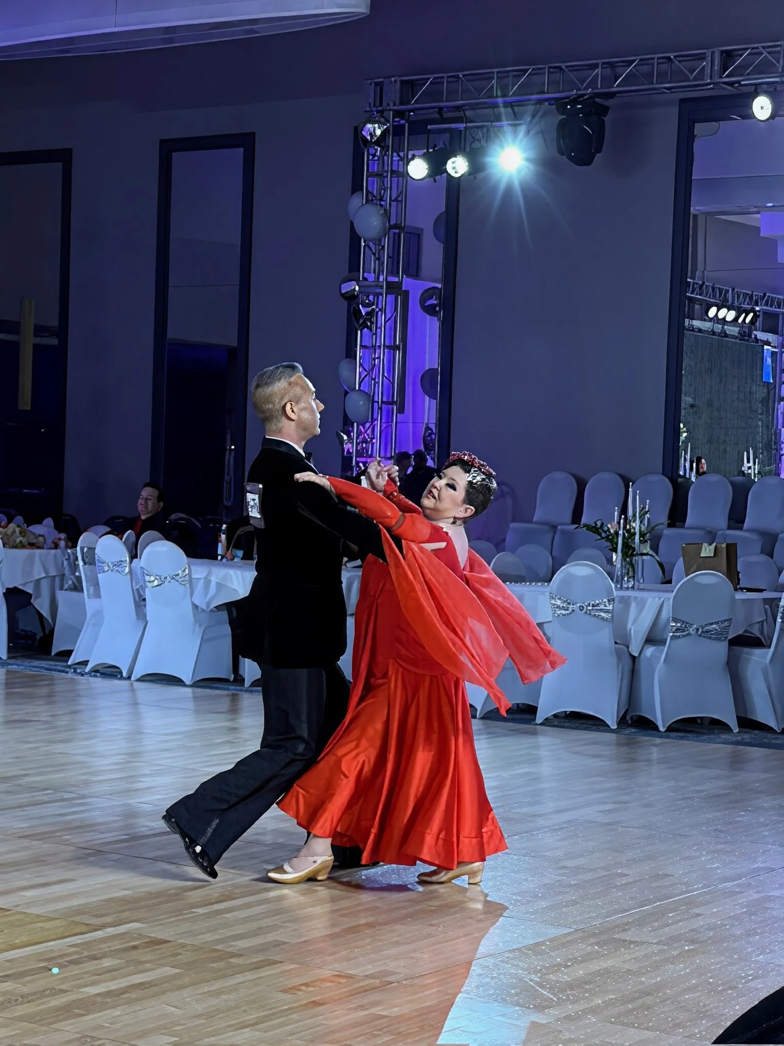 Ballroom dance partners perform on a dance floor, with the woman in a flowing red dress leaning back in frame with her partner.