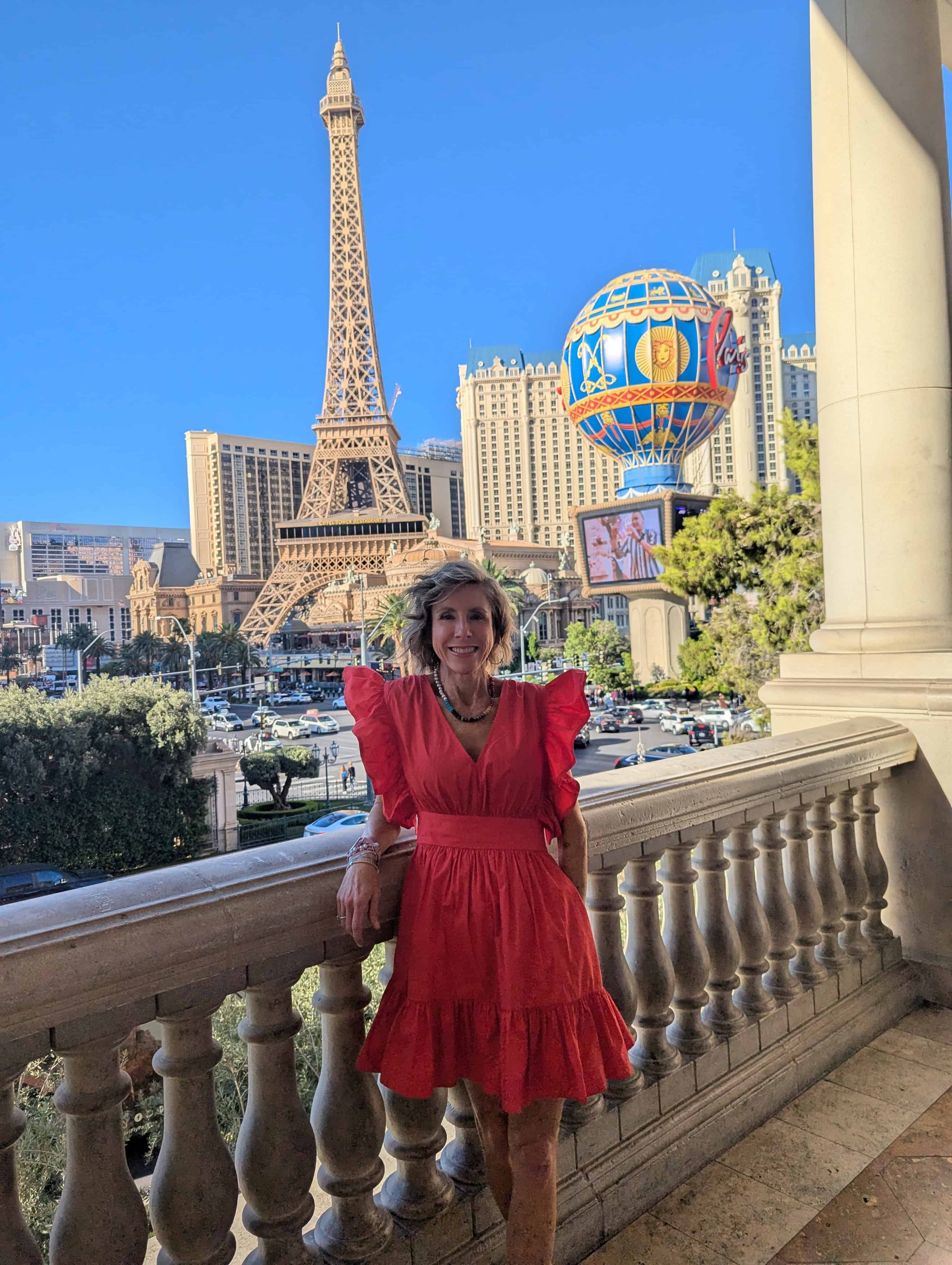 Katie Jensen stands on a balcony in a red dress overlooking the Las Vegas Strip, with the Eiffel Tower replica and Paris Las Vegas in the background.