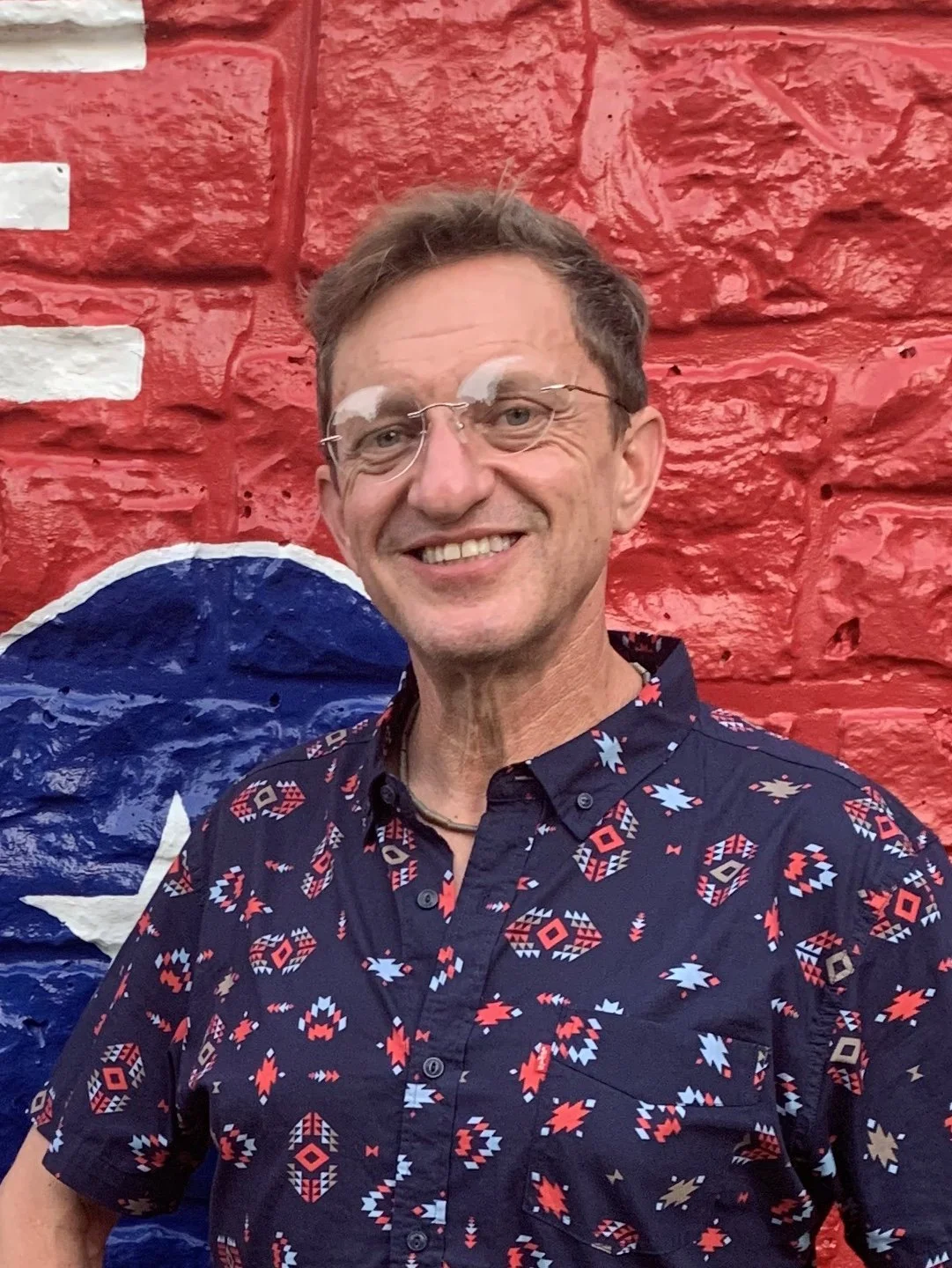 Smiling man in glasses standing in front of a colorful red brick mural wearing a patterned navy button-up shirt.
