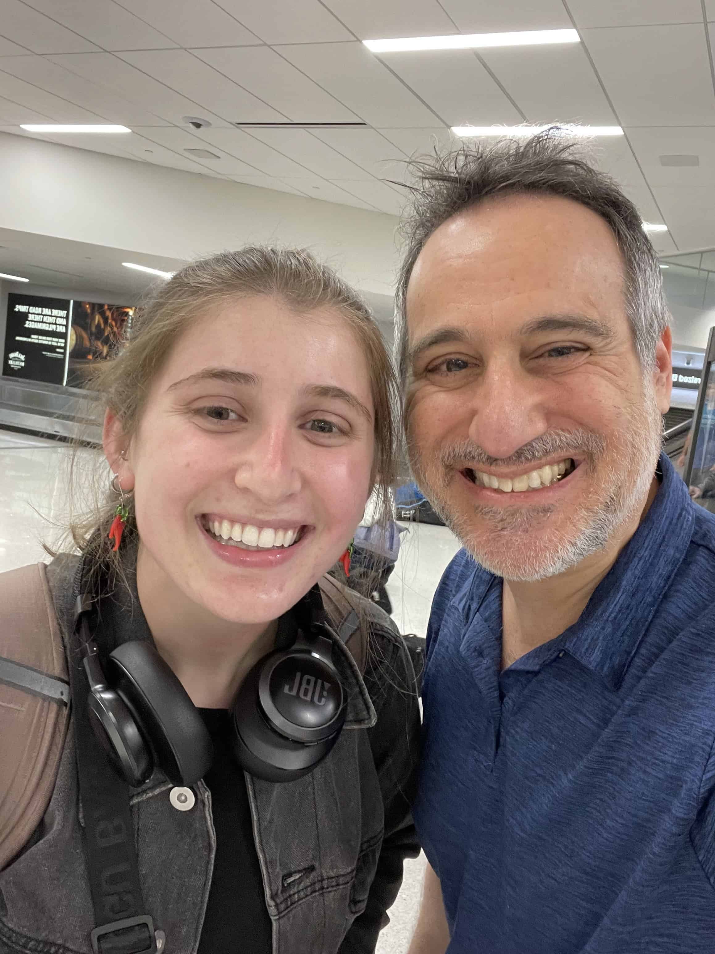 Father and daughter smiling for a selfie in an airport, with the daughter wearing headphones around her neck.
