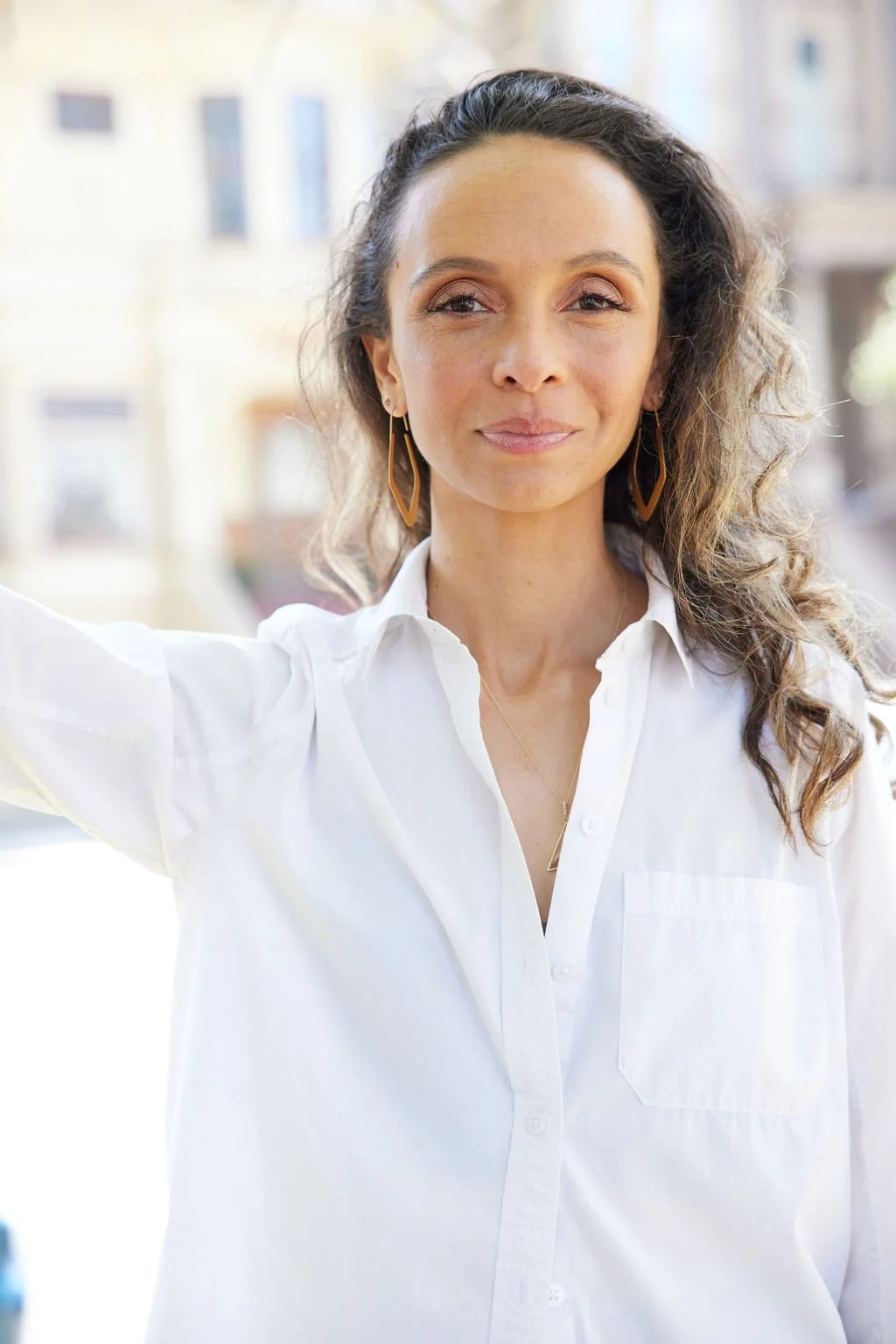 Close-up portrait of Oliveyah Fisch outdoors, wearing a white button-down shirt and long geometric earrings. She looks directly at the camera with a calm, neutral expression against a softly blurred city background.