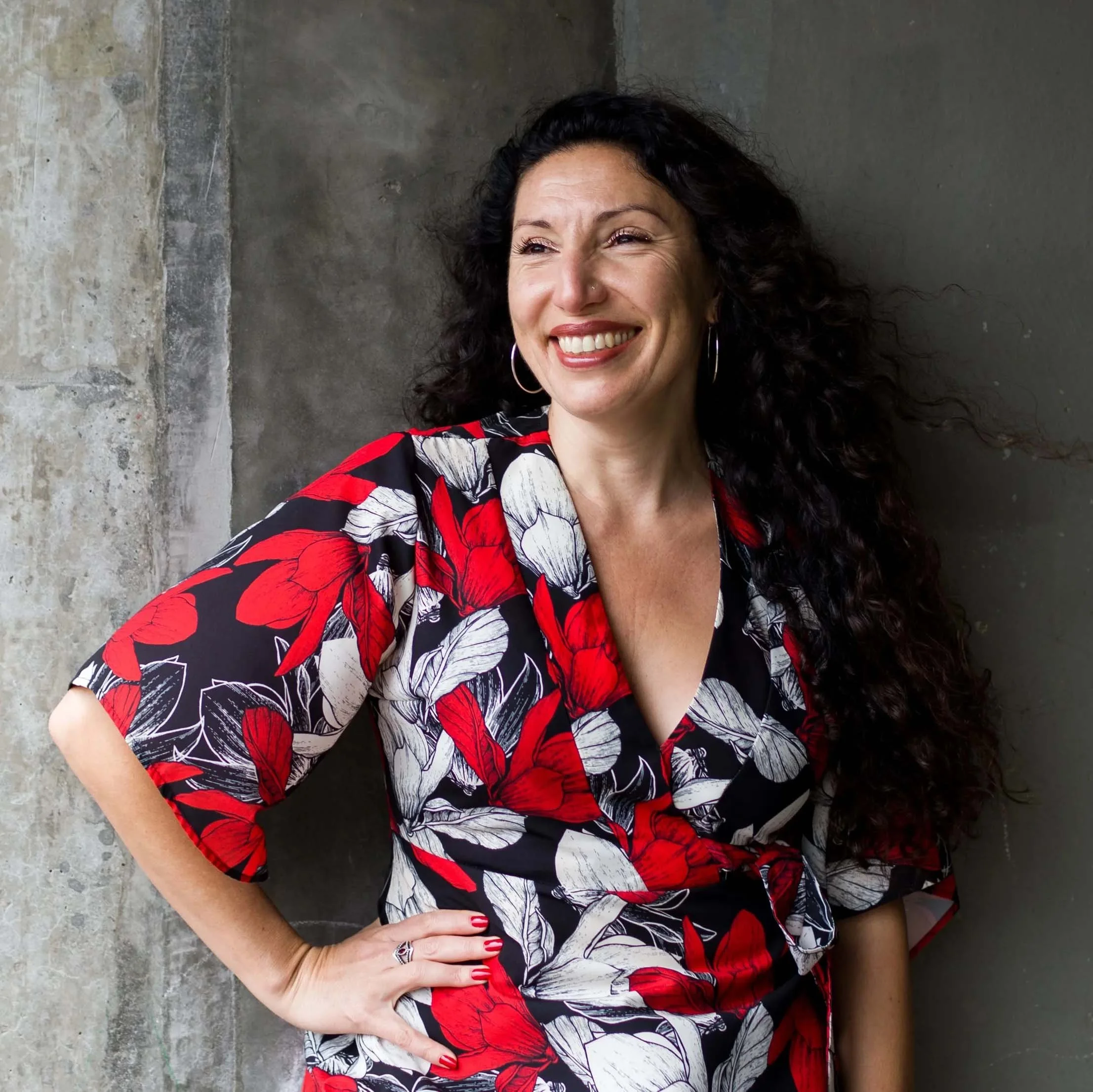 Maritza Schafer smiles while standing with one hand on her hip, wearing a red, black, and white floral dress against a dark wall.